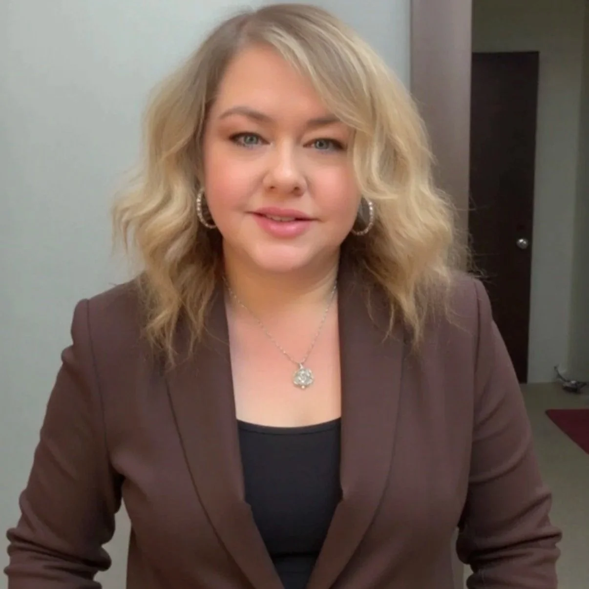 A woman with blonde wavy hair, wearing a brown blazer, black top, silver jewelry, and hoop earrings, standing in an indoor setting.