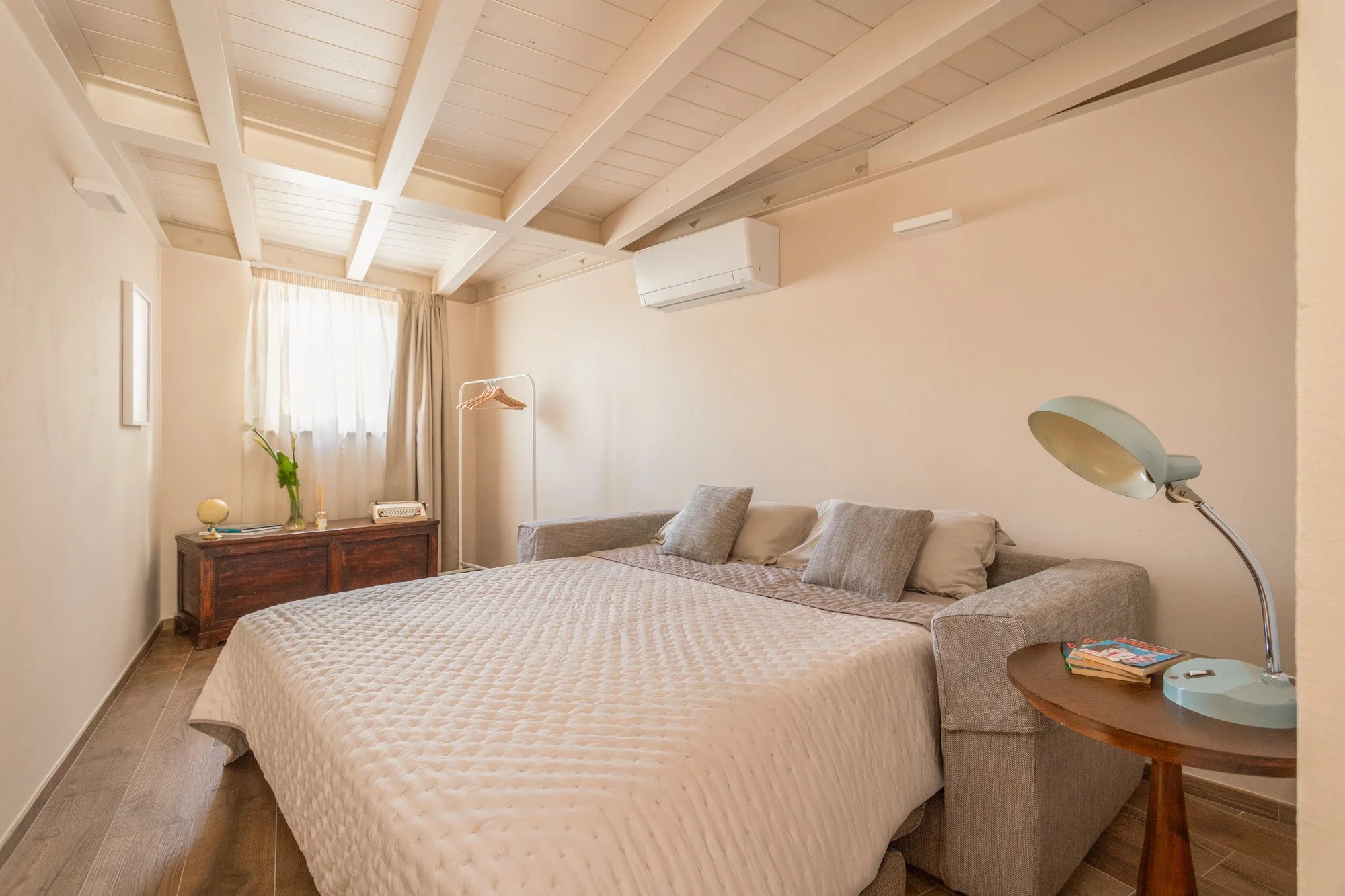 Camera da letto con pareti beige, letto matrimoniale con copriletto beige, cuscini grigi, lampada da tavolo azzurra, finestra con tende chiare, mobile in legno scuro, armadio aperto con spalliera, soffitto con travi, aria condizionata, oggetti decorativi.