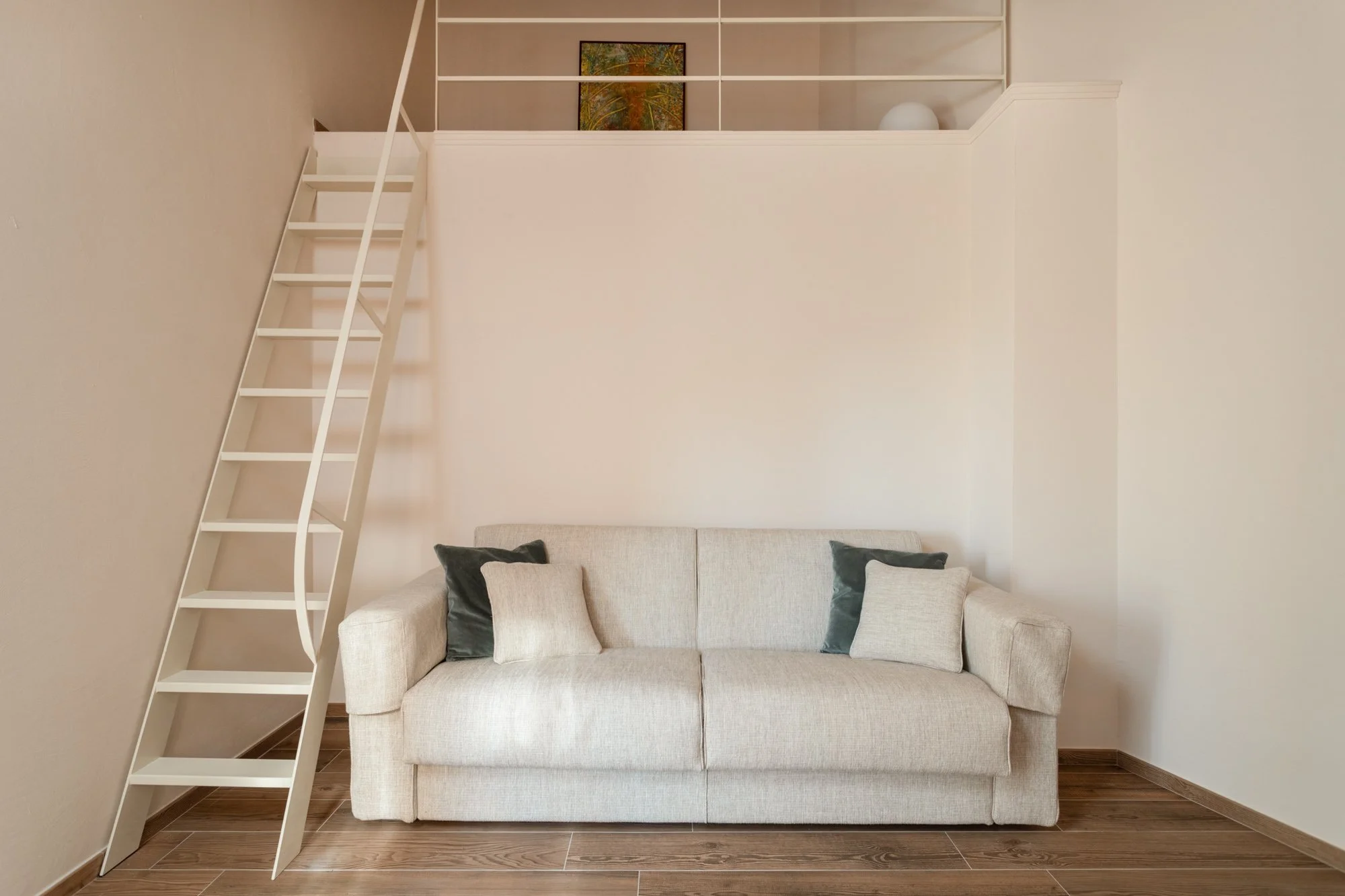 Soggiorno con divano beige e cuscini scuri,scala bianca che conduce a un soppalco, pareti bianche, pavimento in legno.