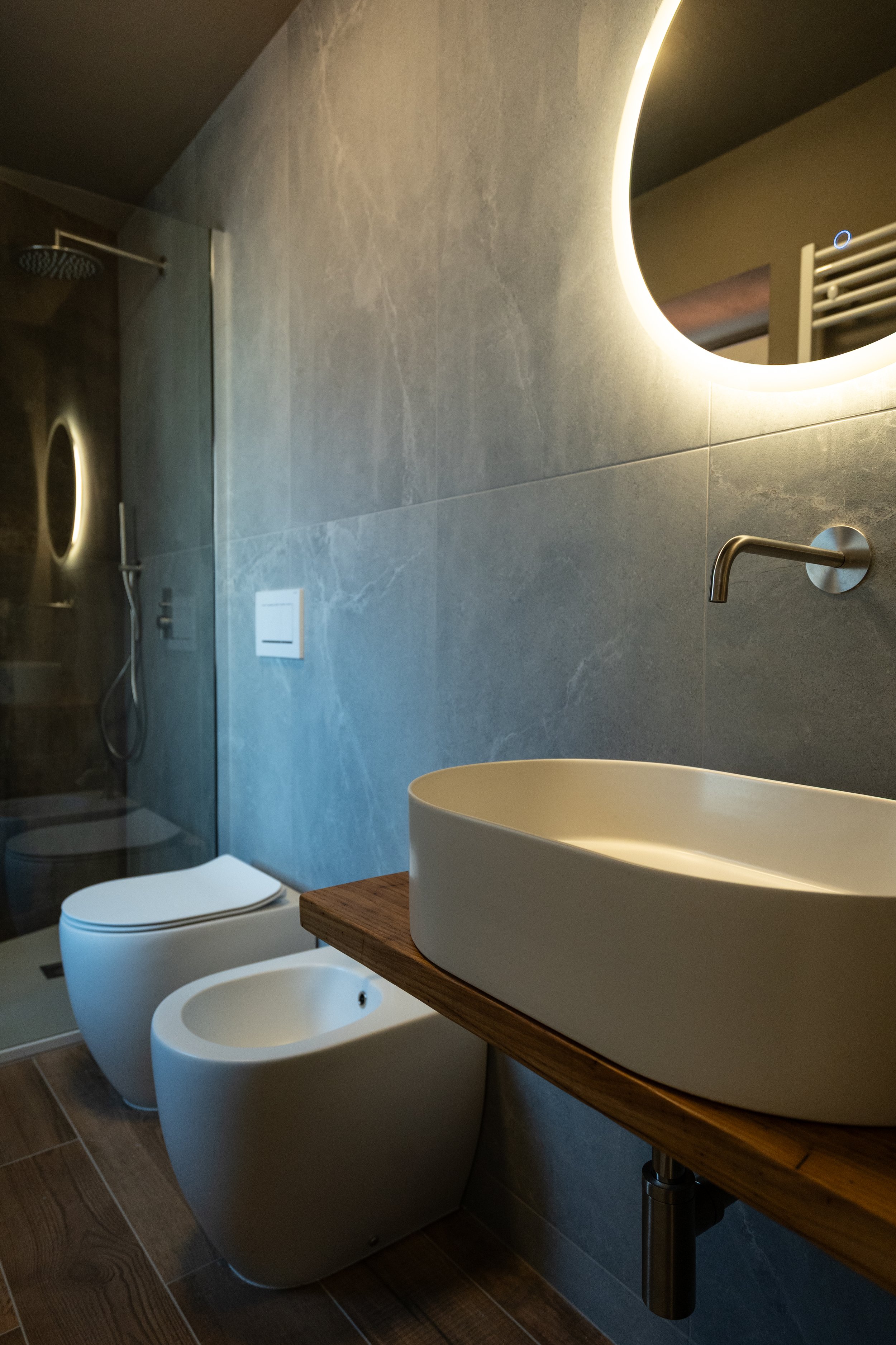 Bagno moderno con lavabo rotondo e specchio luminoso, wc e bidet bianco, in soggiorno con pareti in pietra grigia e pavimento in legno. - Residenze Volpigiano