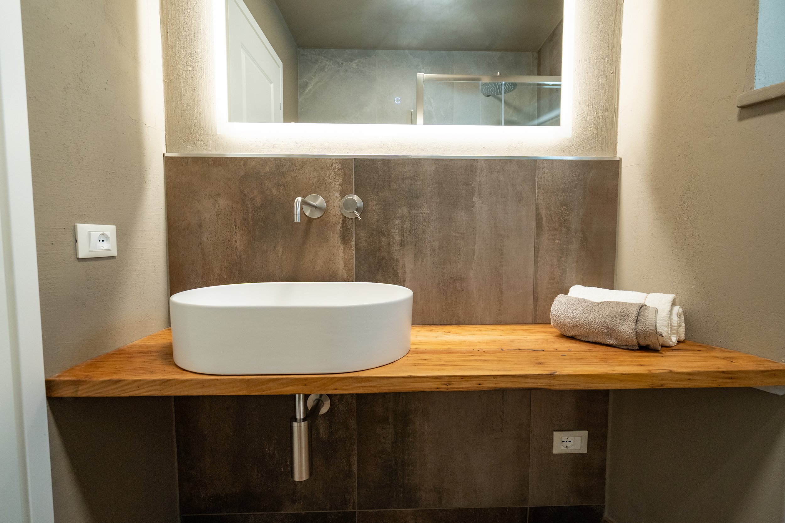 Bagno con lavabo rotondo bianco su piano di legno, specchio con luce, e asciugamani grigi e bianchi su destra. - Residenze Volpigiano