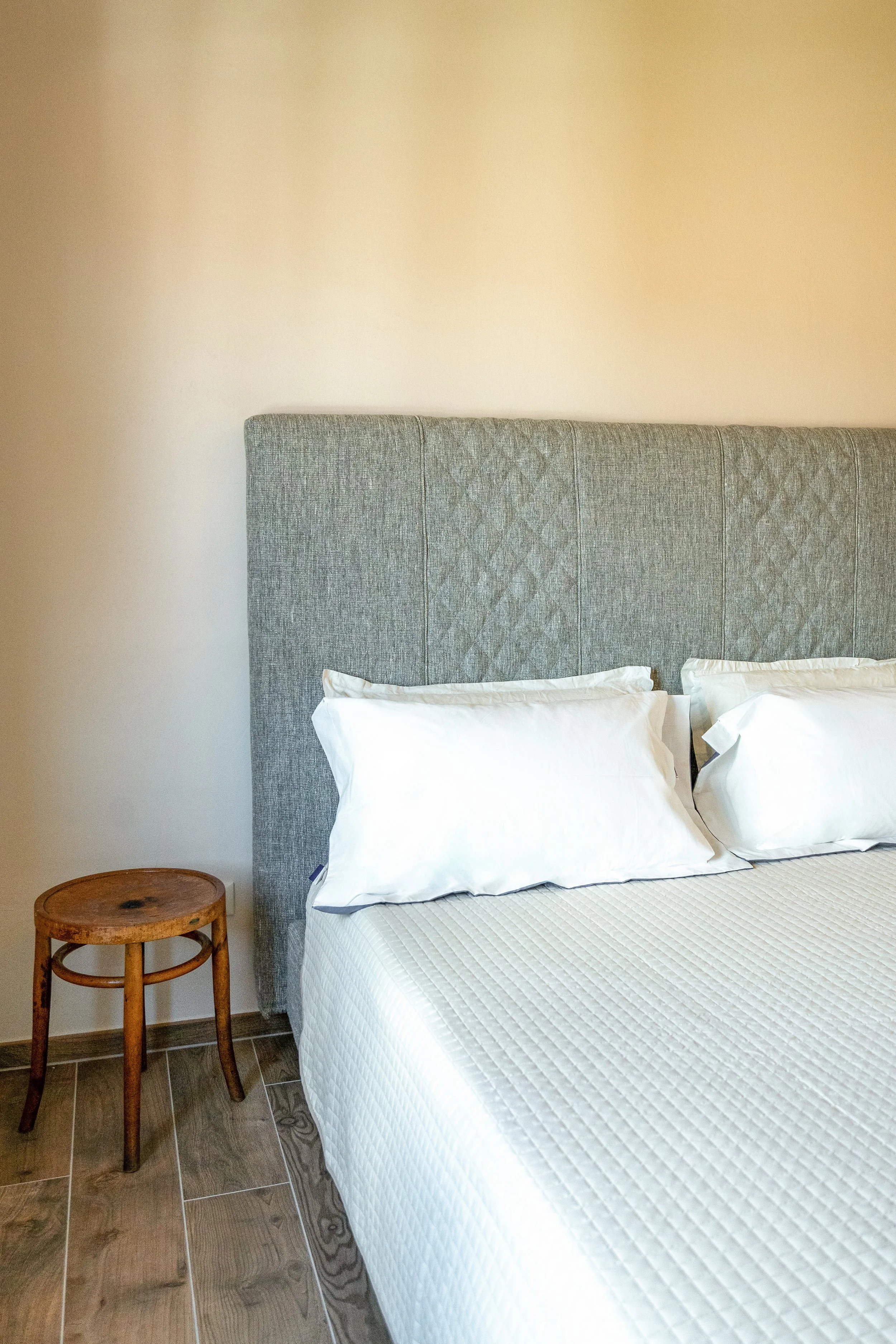 Letto matrimoniale con testiera grigia e due cuscini bianchi, a sinistra un comodino in legno, in una stanza con pareti beige e flooring in legno. - Residenze Volpigiano