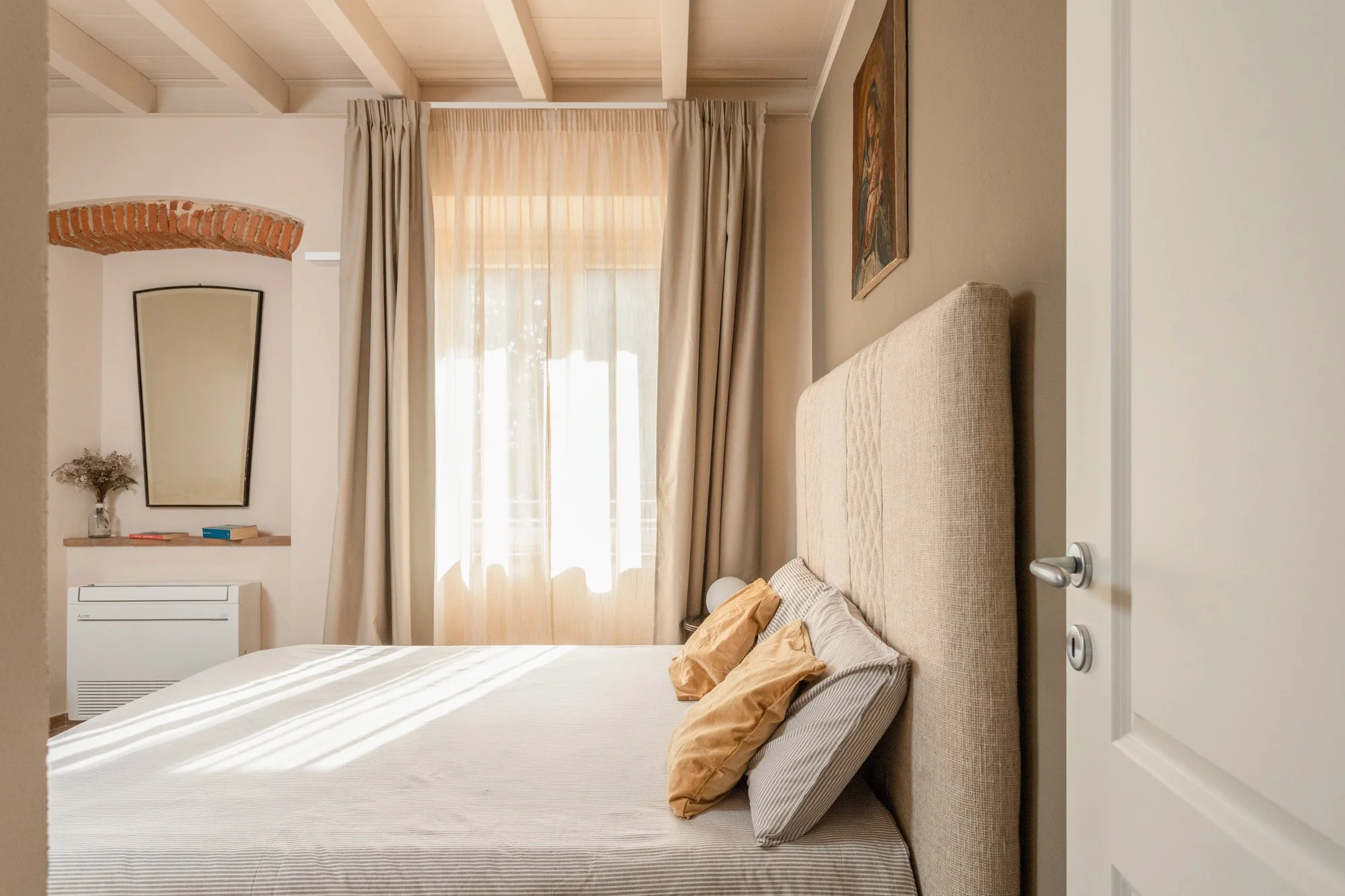 Camera da letto luminosa con letto matrimoniale, grandi finestre con tende chiare, copriletto beige, testiera imbottita, decorazioni alle pareti e uno specchio sulla parete di fronte