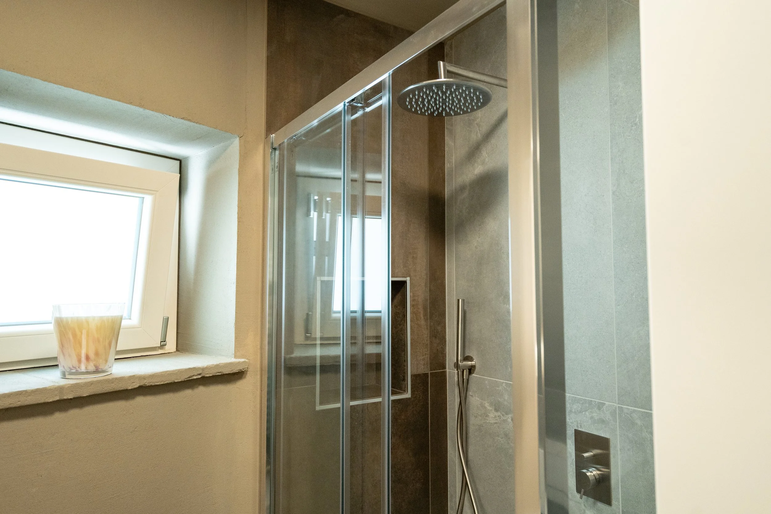 Doccia moderna con pioggia, all'interno di un bagno con pareti in piastrelle grigie, accanto a una finestra con una candela sulla finestra. - Residenze Volpigiano