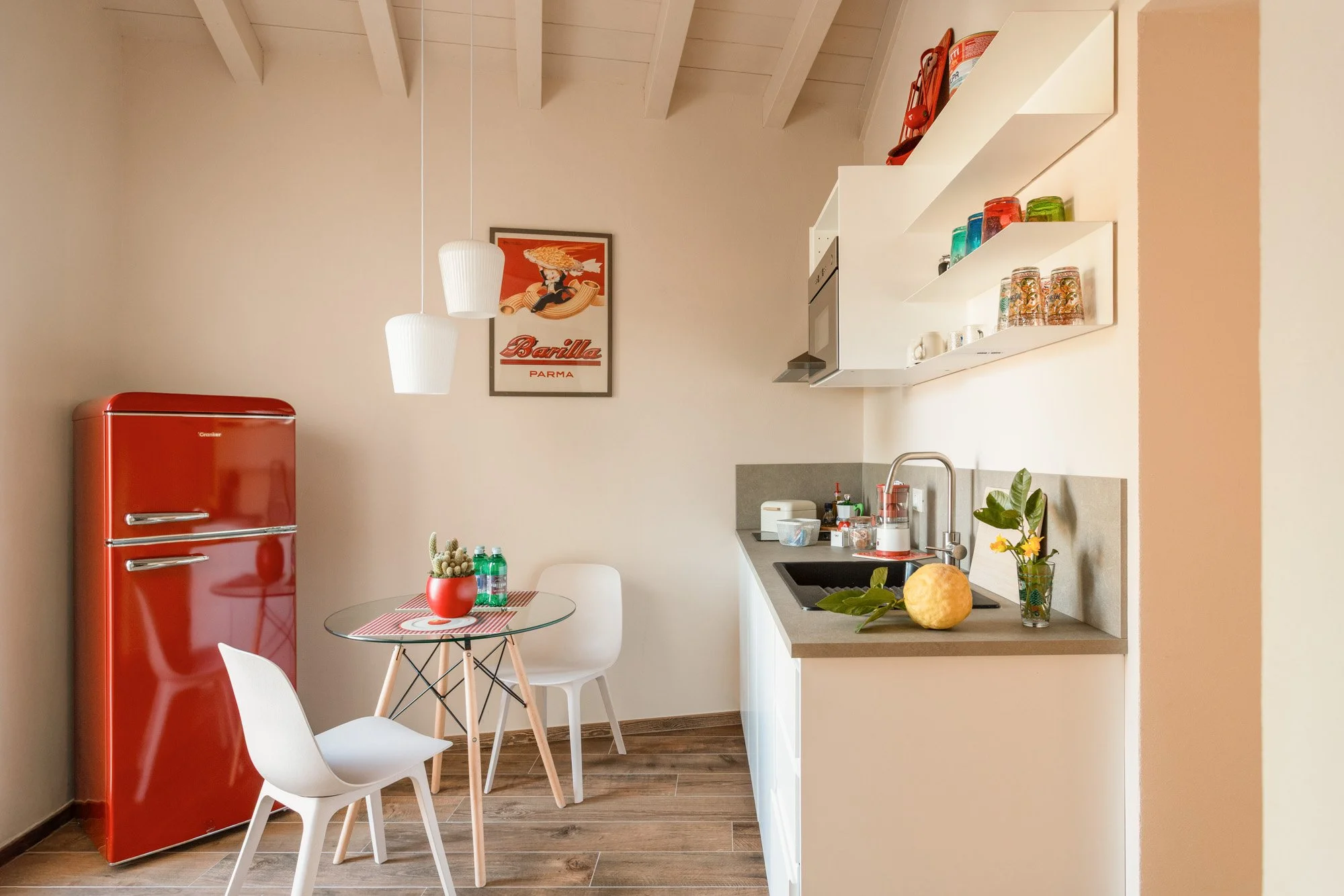 Cucina minimalista con frigorifero rosso, tavolo rotondo con due sedie bianche, decorazioni verdi e pareti chiare. Per Kura