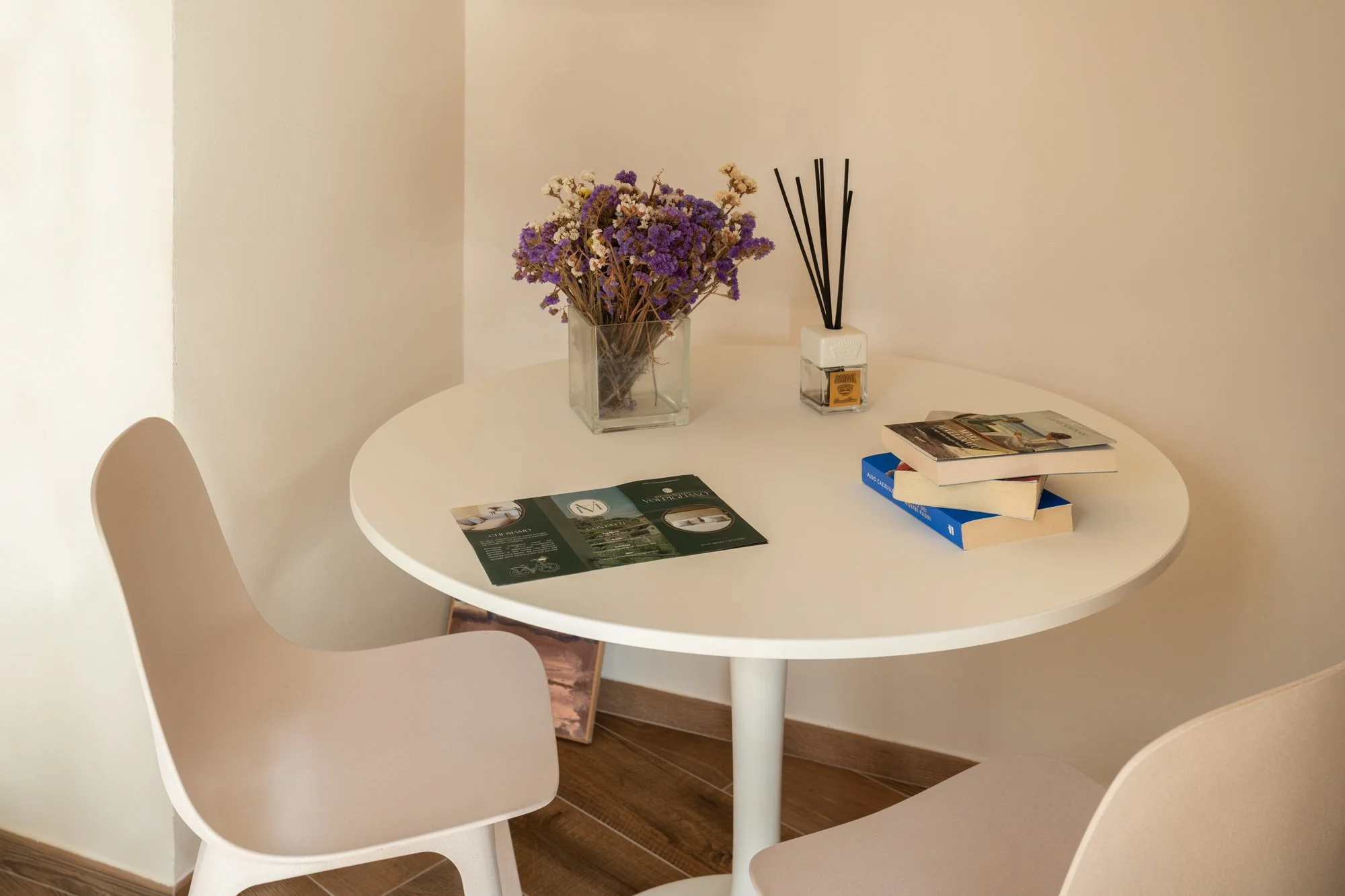 Tavolo rotondo bianco con due sedie beige, decorato con un vaso di fiori secchi, un diffusore di aromi e alcuni libri e volantini.