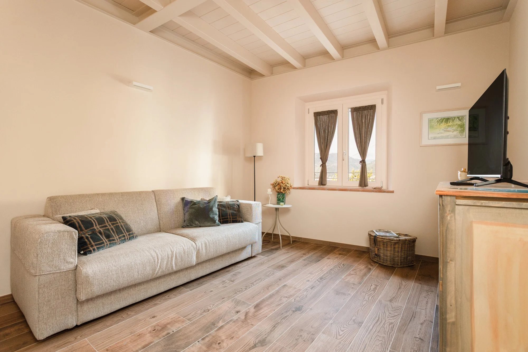 Soggiorno con divano beige, tavolino con vasi di fiori, televisore su mobile in legno, finestra con tende marroni, pavimento in parquet.