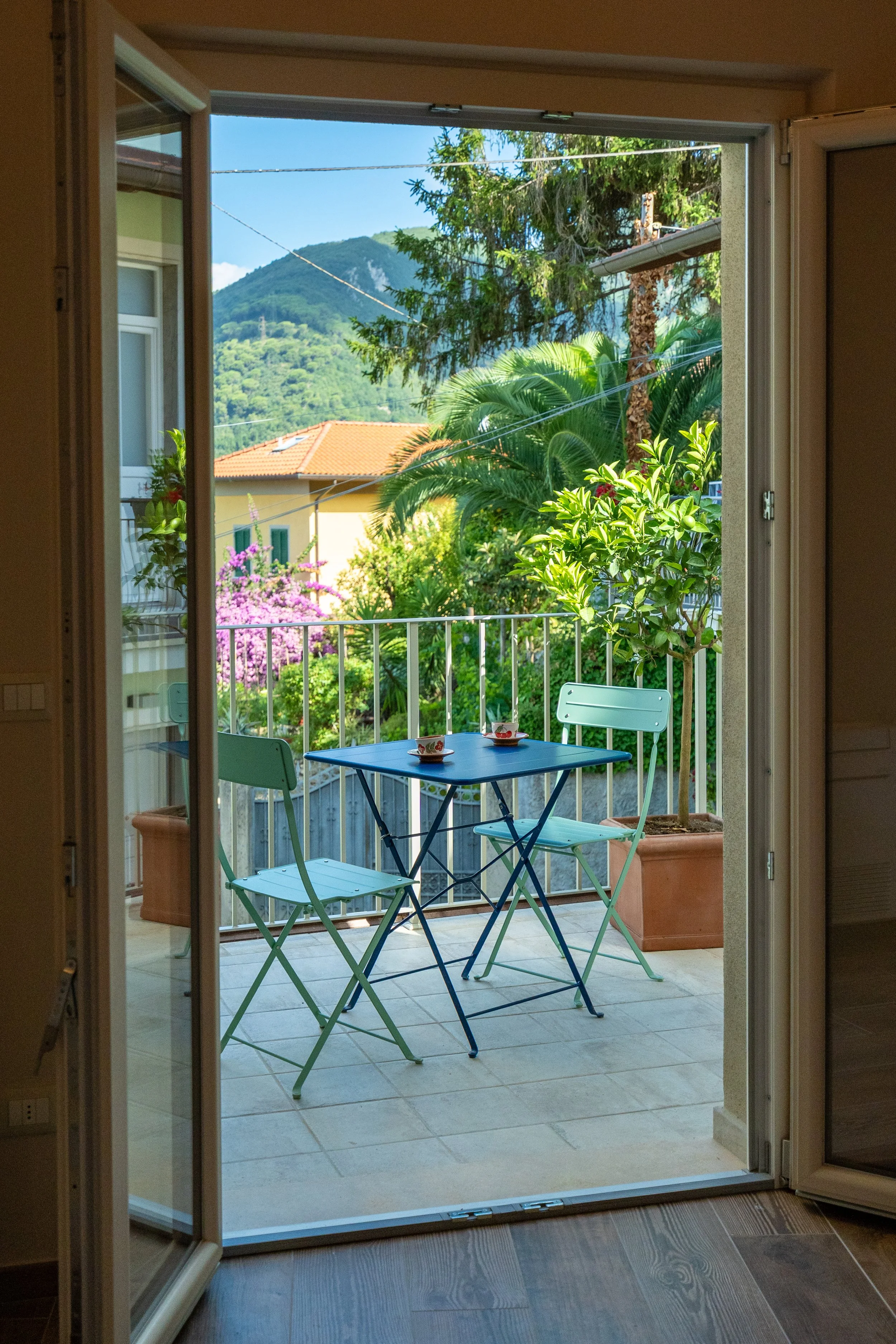 Vista di un balcone con tavolo e sedie azzurre, piante verdi e un albero, con montagne sullo sfondo e cielo azzurro. - Residenze Volpigiano