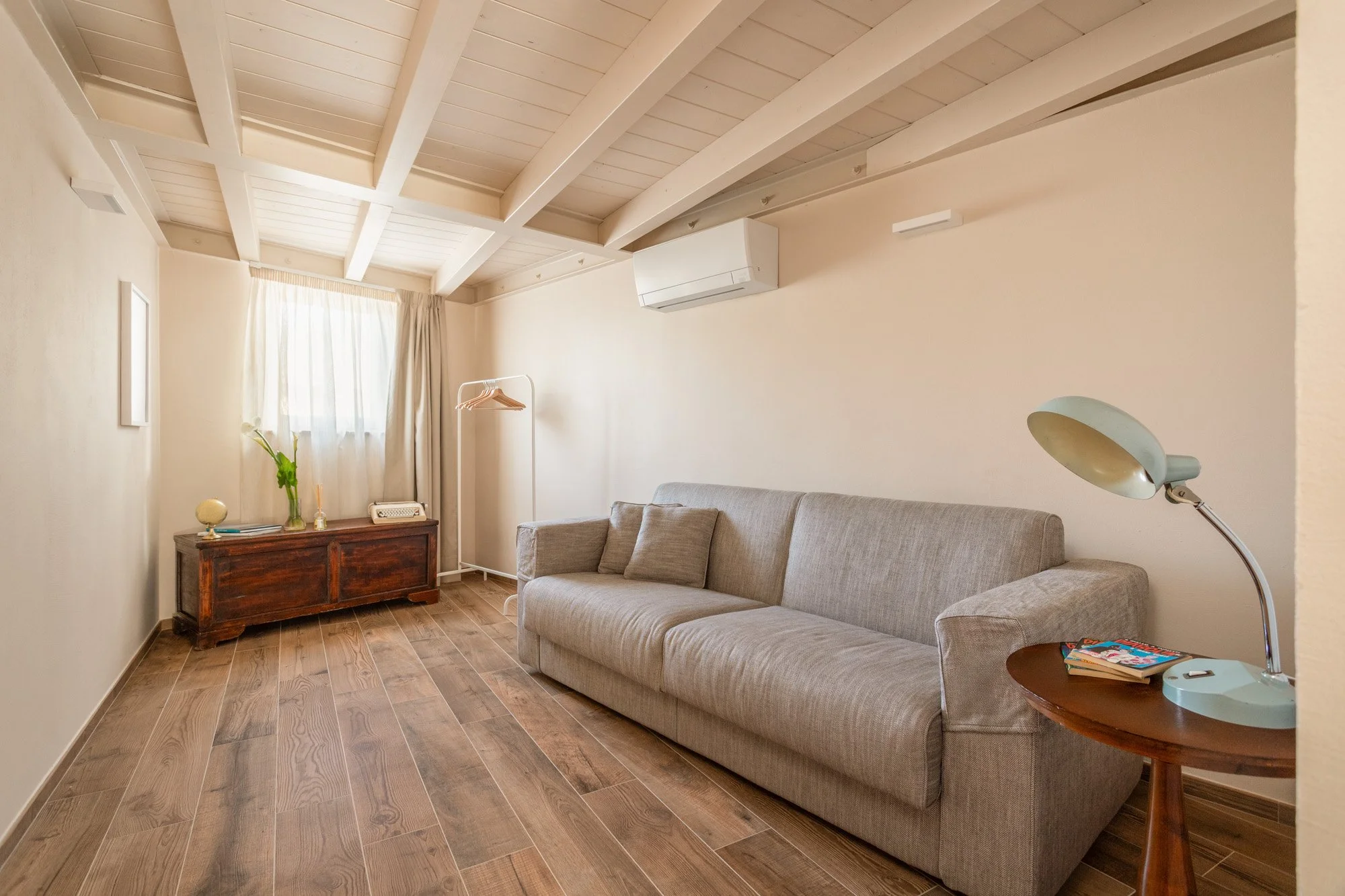 Salotto minimalista con divano grigio, lampada da tavolo, tavolino in legno, armadio antico, e finestra con tende leggere.