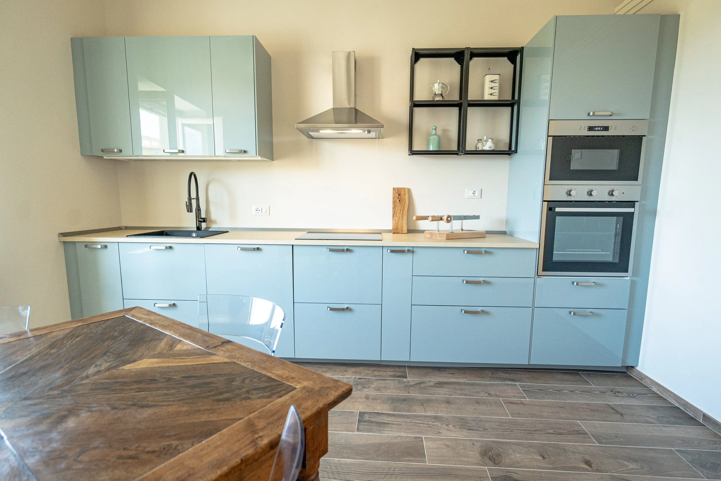 Cucina moderna con mobili blu e pareti chiare, include forno, microonde, cappa, lavello e un tavolo in legno chiaro. - Residenze Volpigiano