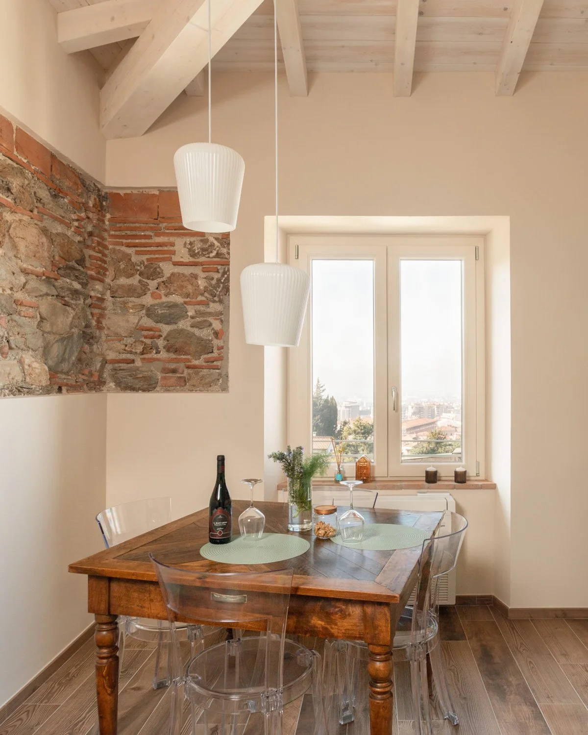 Interno di una stanza con tavolo in legno, sedie trasparenti, due lampade pendenti bianche, vaso con piante, vista cittadina dalla finestra. Pareti in pietra e intonaco, soffitto con travi in legno