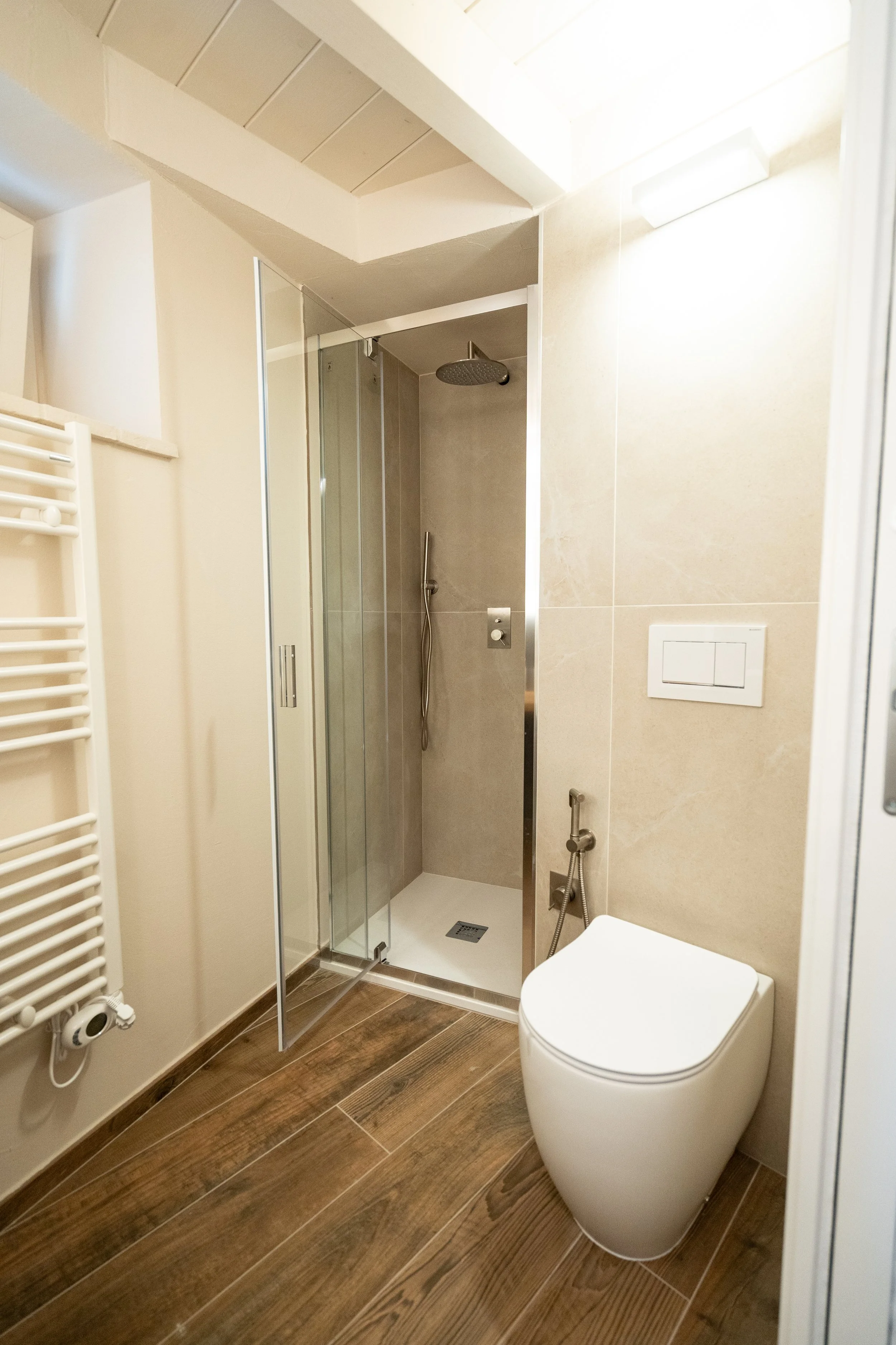 Bagno con doccia a vetri, WC moderno, rivestimenti in tonalità neutre e pavimento in legno.  - Residenze Volpigiano