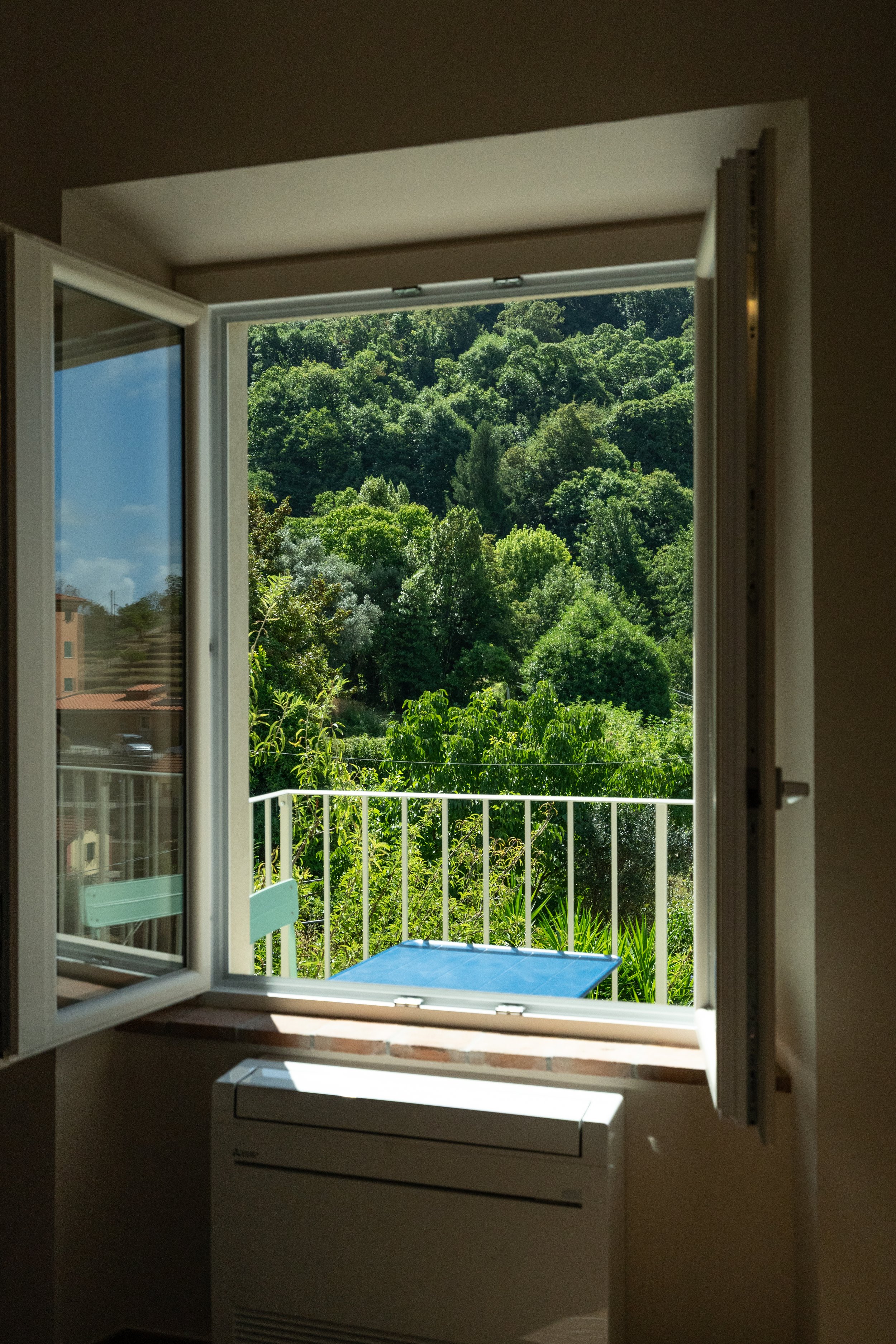 Finestra aperta che si affaccia su un balconcino con vista su un bosco verde e folto, con un tavolo blu fuori e un condizionatore sotto la finestra - Residenze Volpigiano