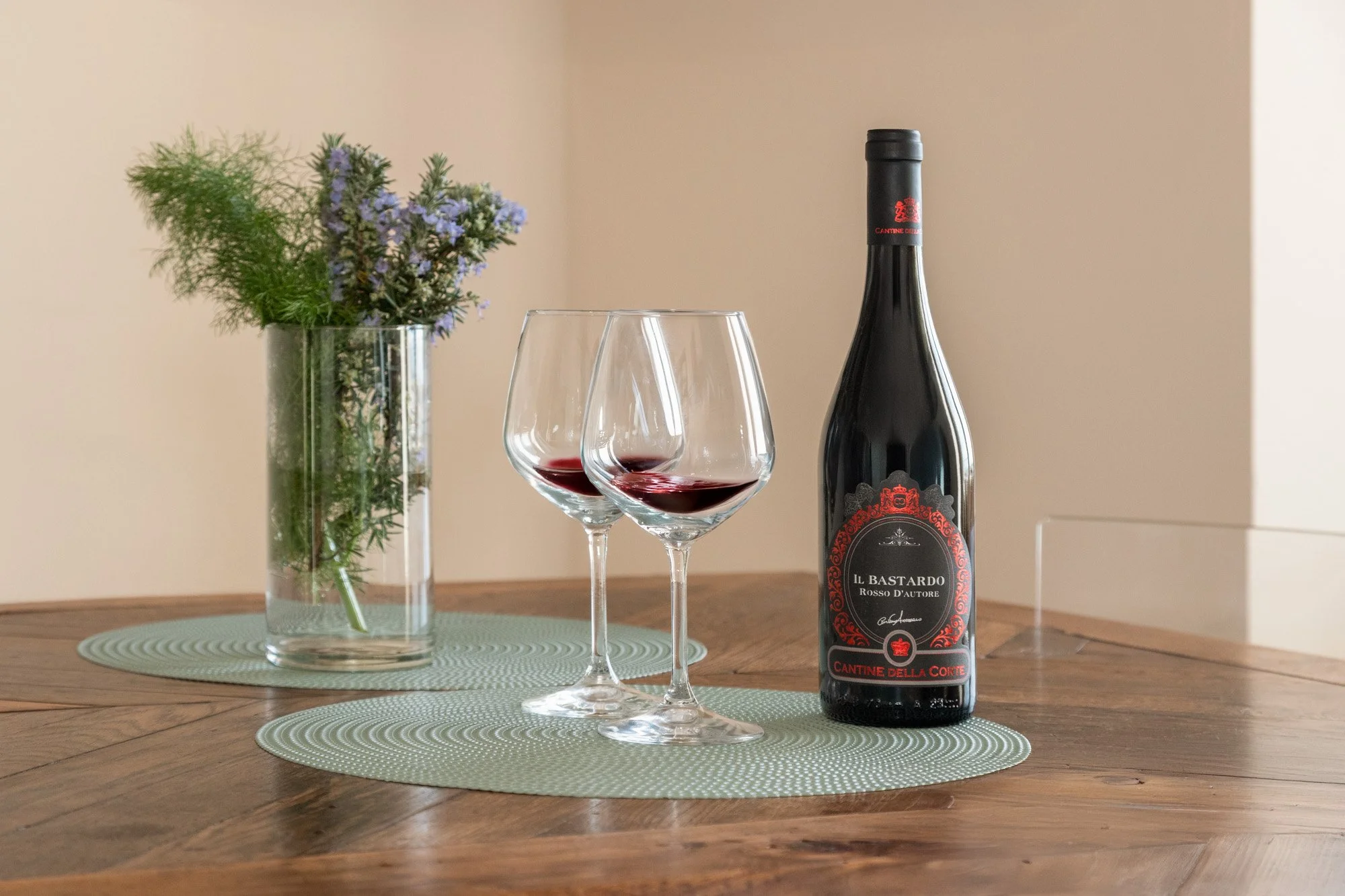 Bottiglia di vino rosso, due bicchieri di vino rovesciati con poco vino, vaso con fiori e piatti di vimini su tavolo di legno.