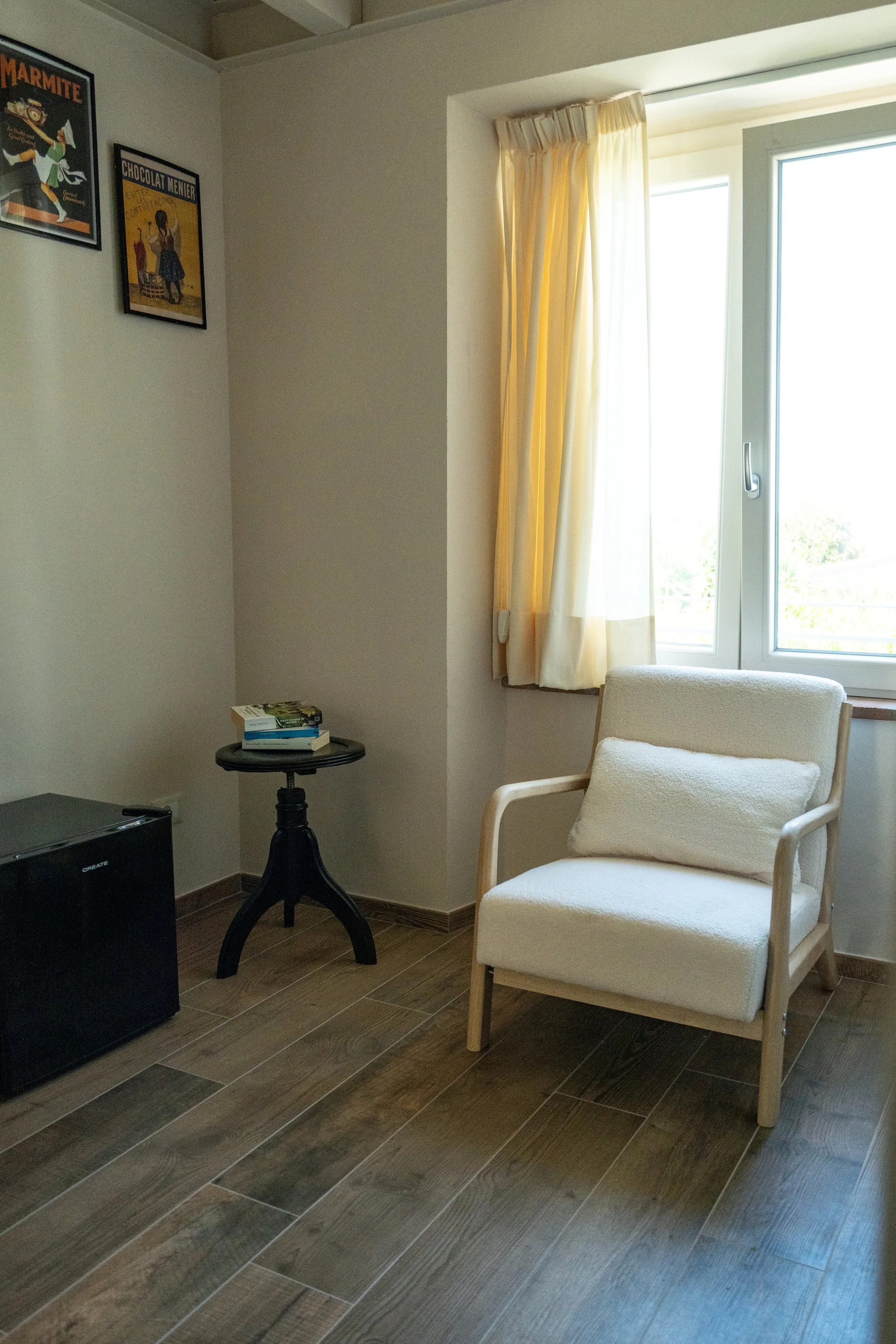 Angolo di una stanza con una poltrona beige con cuscino, una finestra con tenda gialla, un tavolo nero con libri, e una parete con due poster vintage. Pavimento in legno. - Residenze Volpigiano