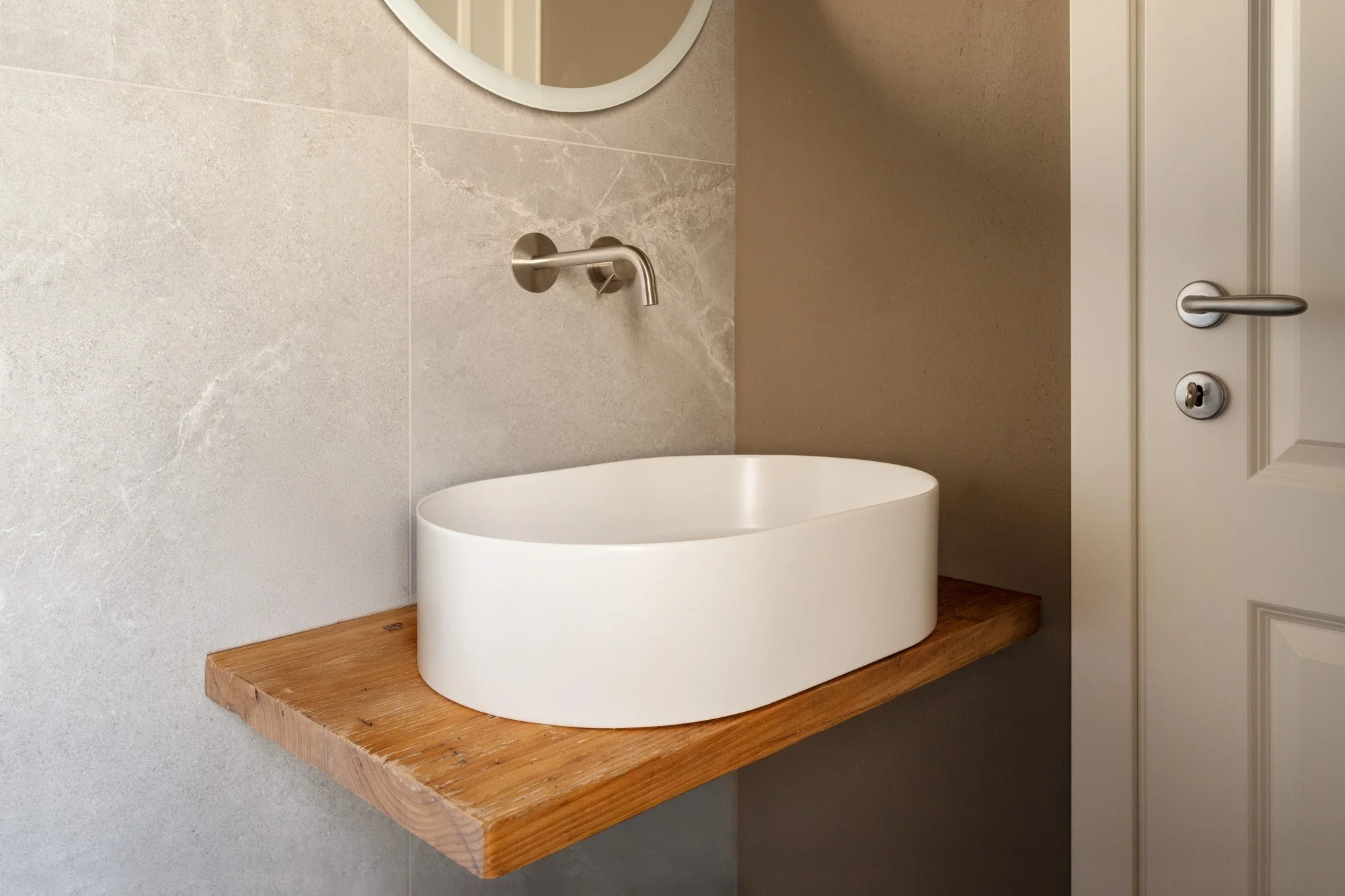 Lavabo bianco rotondo sopra un piano di legno, pareti beige e una porta bianca con maniglia di metallo.
