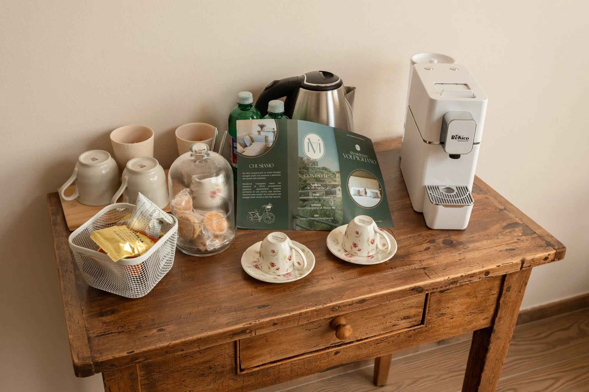 Tavolo di legno con macchina per caffè, tazze, bicchieri, bottiglie d'acqua, brochure e snack.