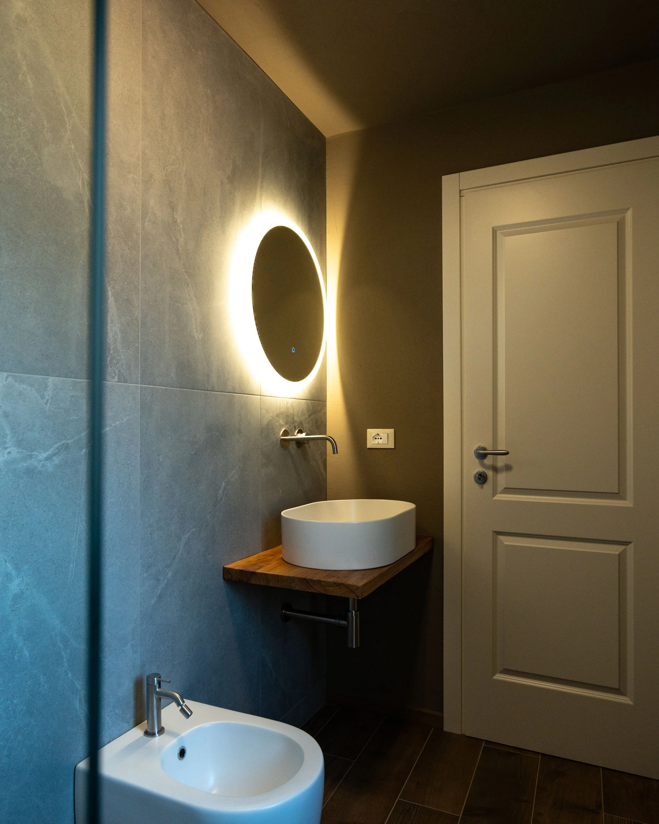 Bagno moderno con lavabo rotondo su piano di legno, specchio rotondo con illuminazione a LED e porta bianca. Arcobaleno di luce intorno allo specchio, muro in piastrelle grigie e pavimento in legno scuro. - Residenze Volpigiano