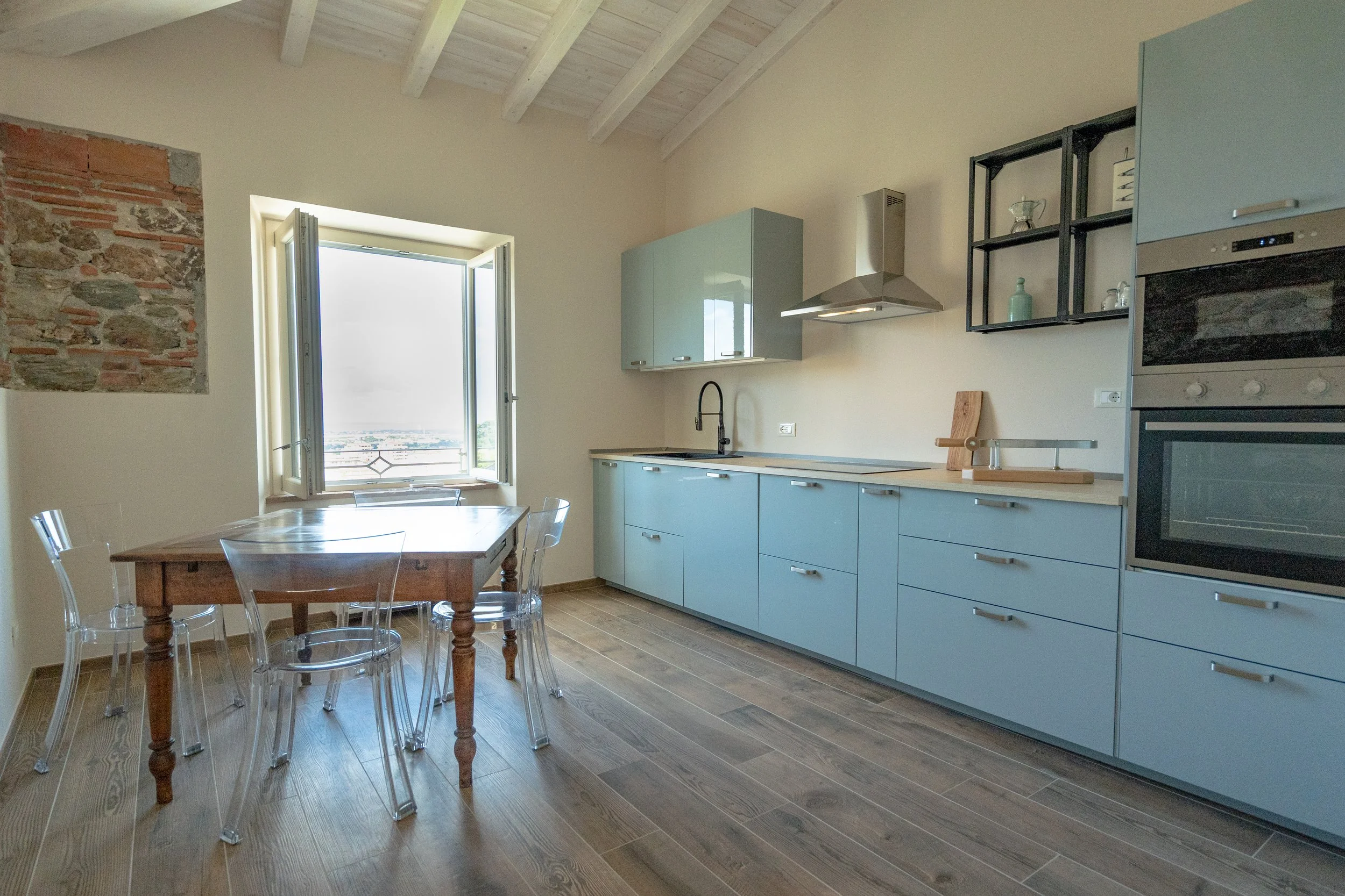 Cucina moderna con pareti neutre, armadietti azzurri, un tavolo di legno con sedie di plastica trasparente e una finestra aperta che mostra un paesaggio. - Residenze Volpigiano
