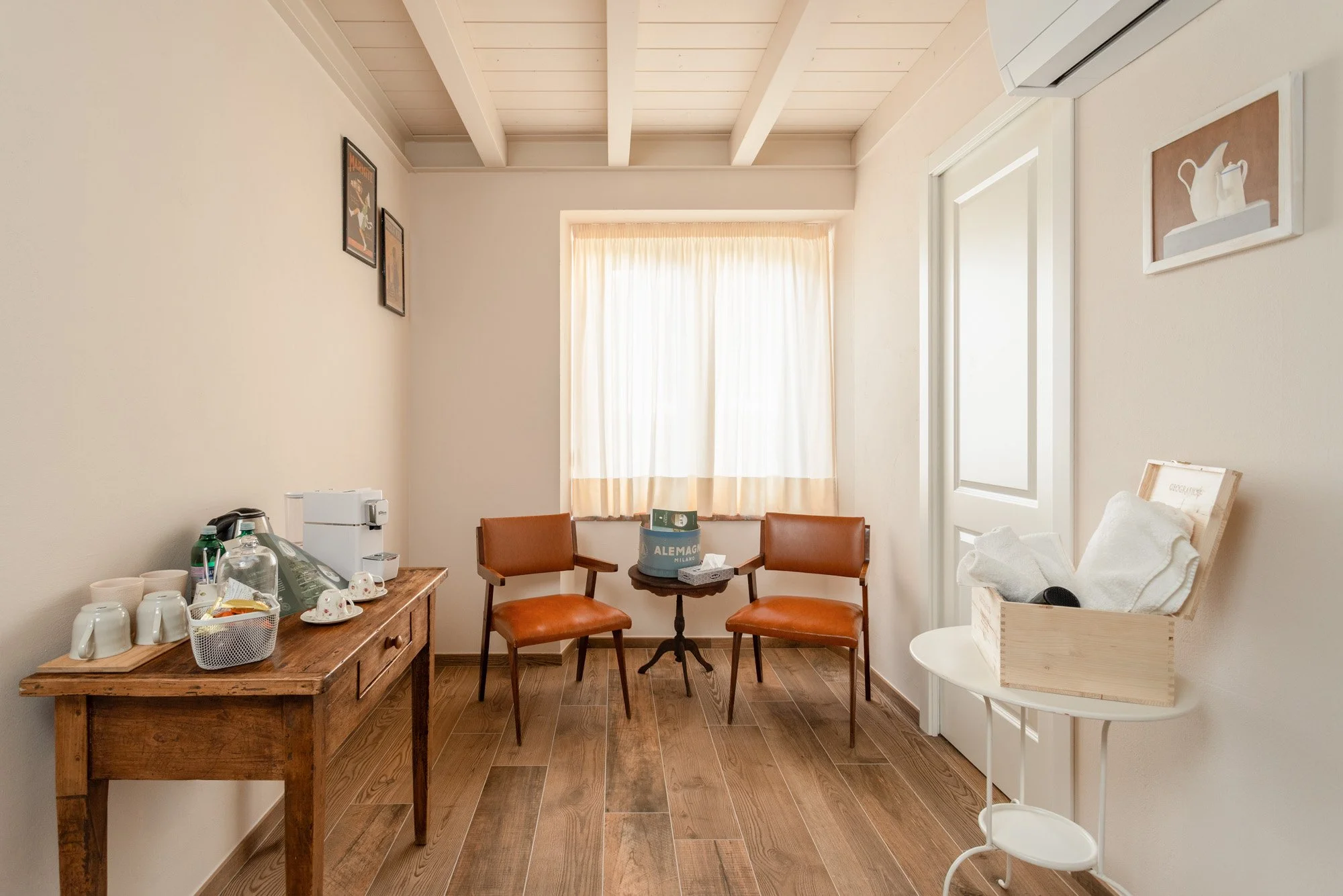 Stanza con due sedie di pelle marrone di fronte a una finestra con tende chiare, tavolo rotondo tra le sedie con scatola e tessuti, pareti decorano pai di quadri, tavolo di legno con macchina per caffè, tazze e bottiglie, angolo con scatola di asciugamani e altri oggetti.