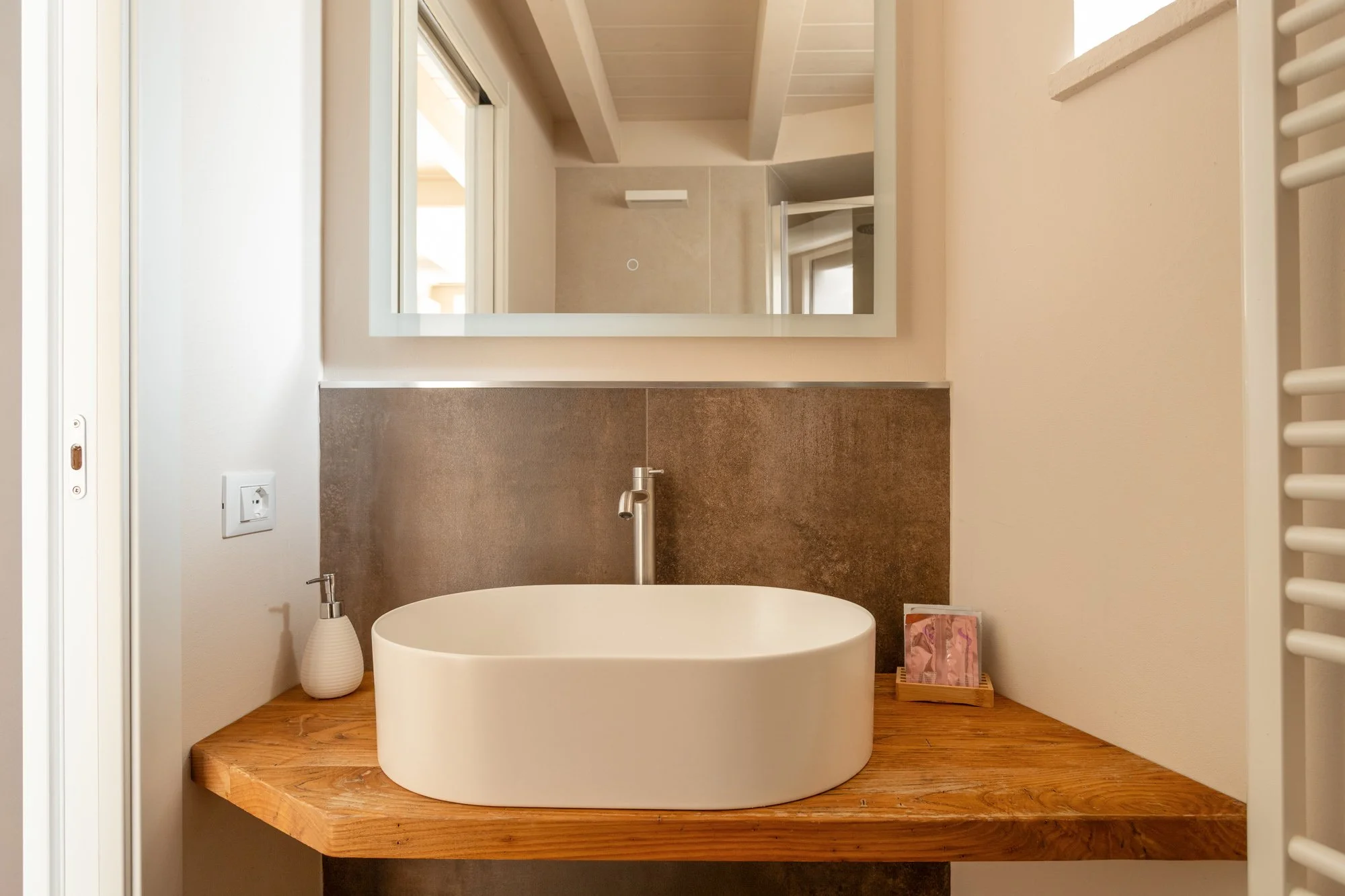 Lavandino rotondo sopra un banco di legno, con uno specchio sopra, pareti beige e una finestra