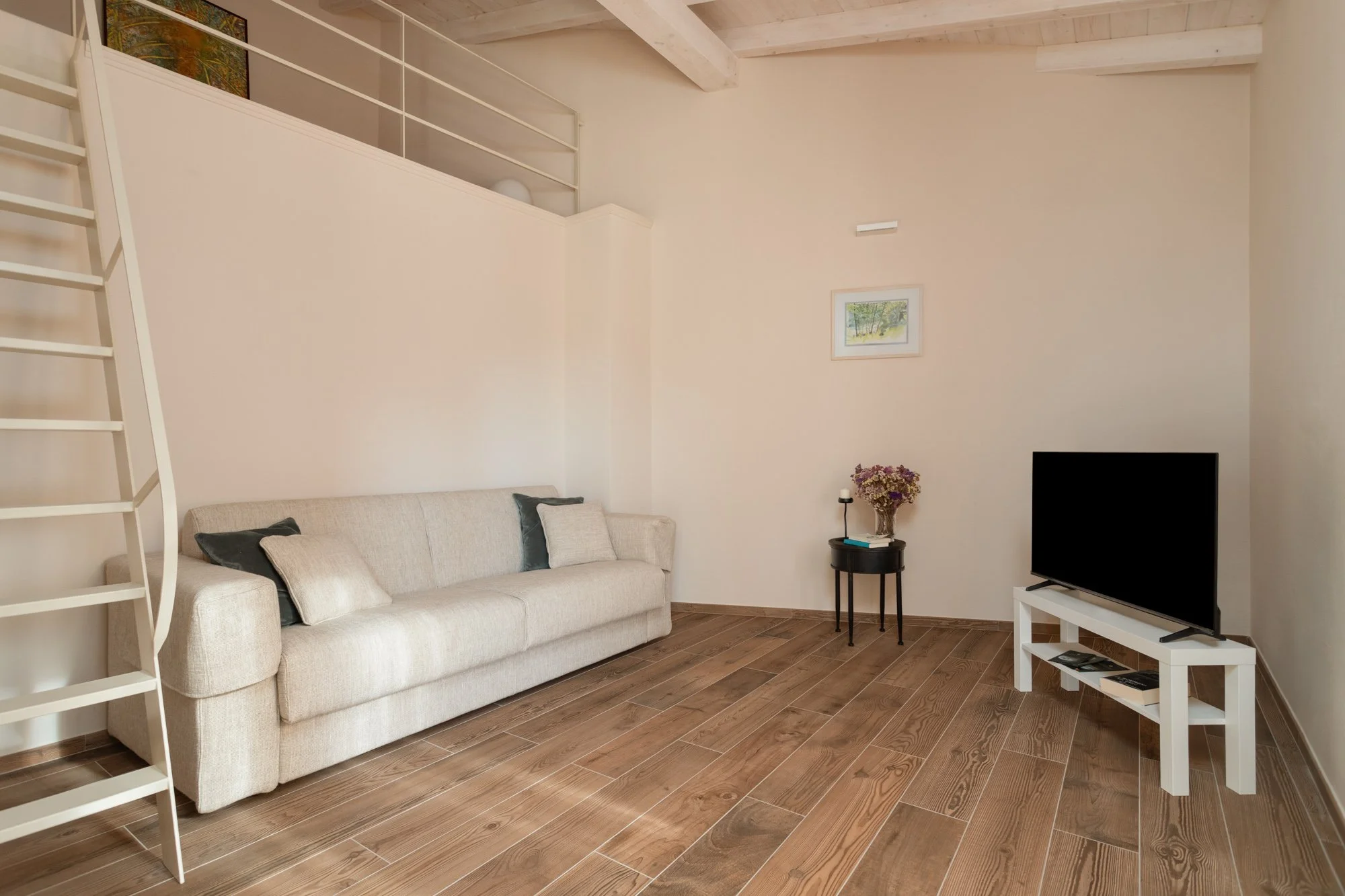 Soggiorno con divano beige, televisore su tavolo bianco, tavolo con bouquet di fiori, quadro e lampada sul muro, rampa di scale e soffitto in legno