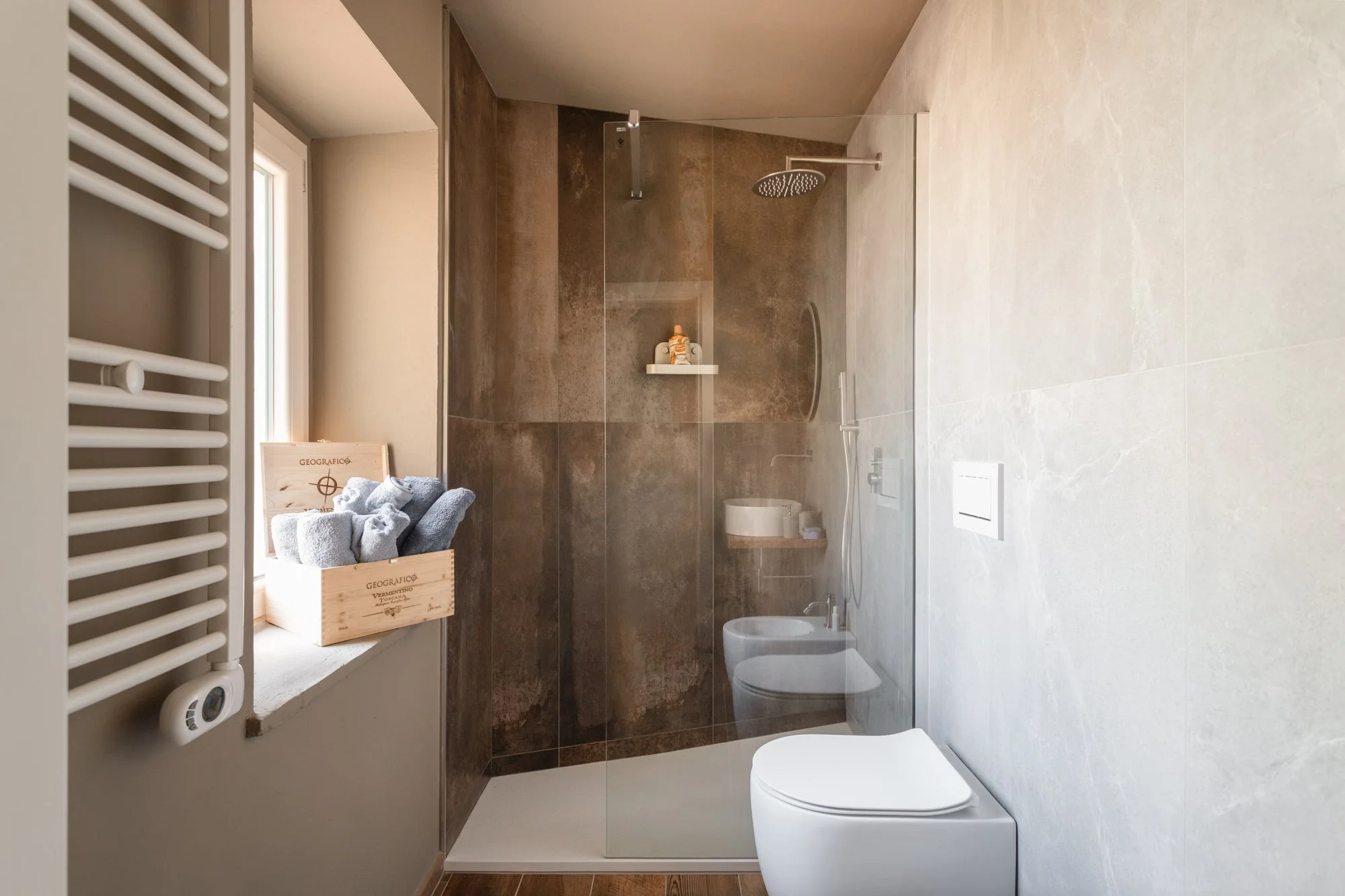 Bagno moderno con pareti in piastrelle marroni e beige, cabina doccia con parete in vetro, scalda asciugamani bianco, finestra con tenda, box con asciugamani grigi e scatola in legno, sanitari bianchi.
