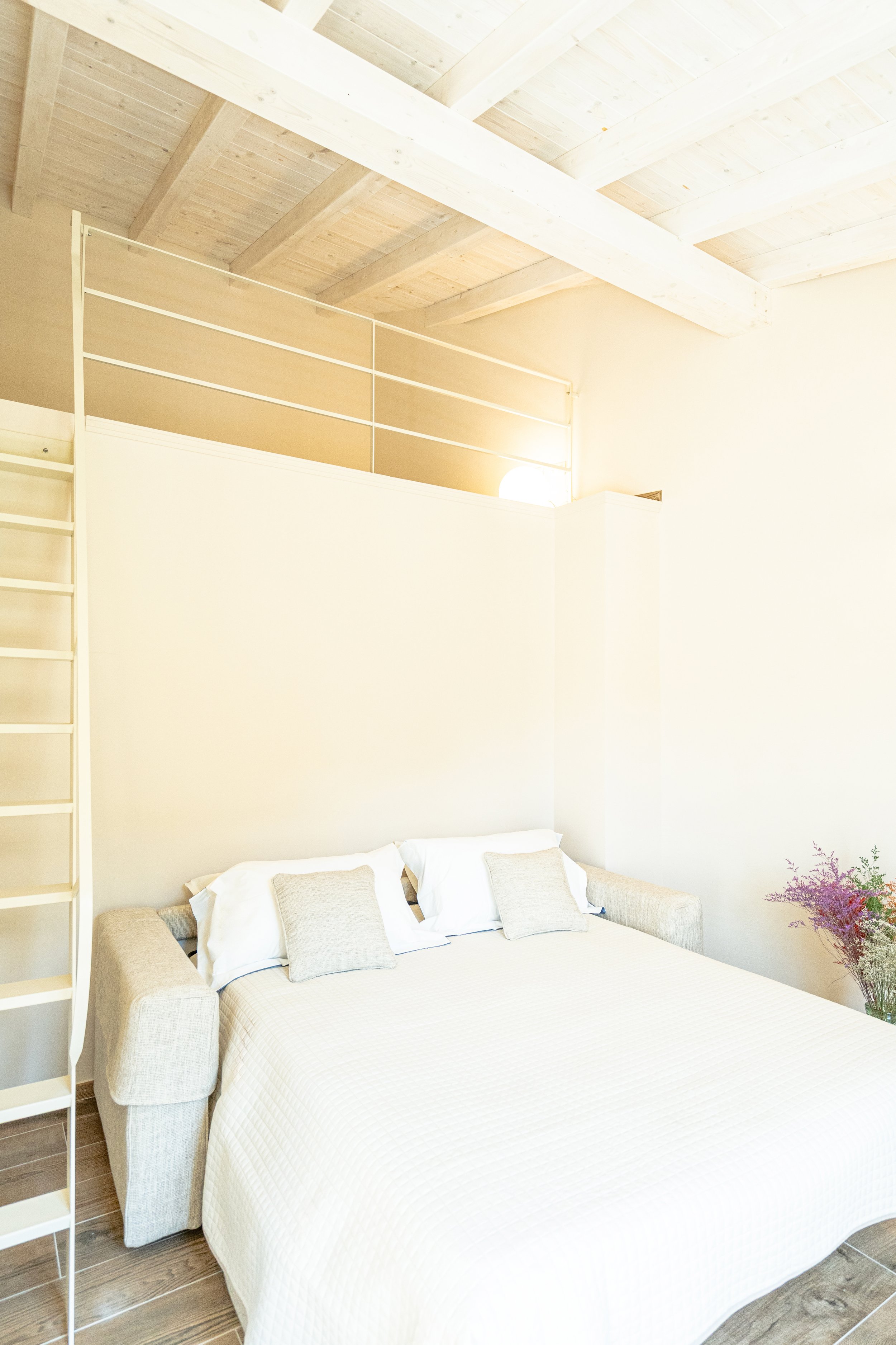 Camera da letto con divano letto, biancheria bianca, cuscini chiari, pareti beige, soffitto con travi in legno e una parete sopra con ringhiera, corretta illuminazione - Residenze Volpigiano