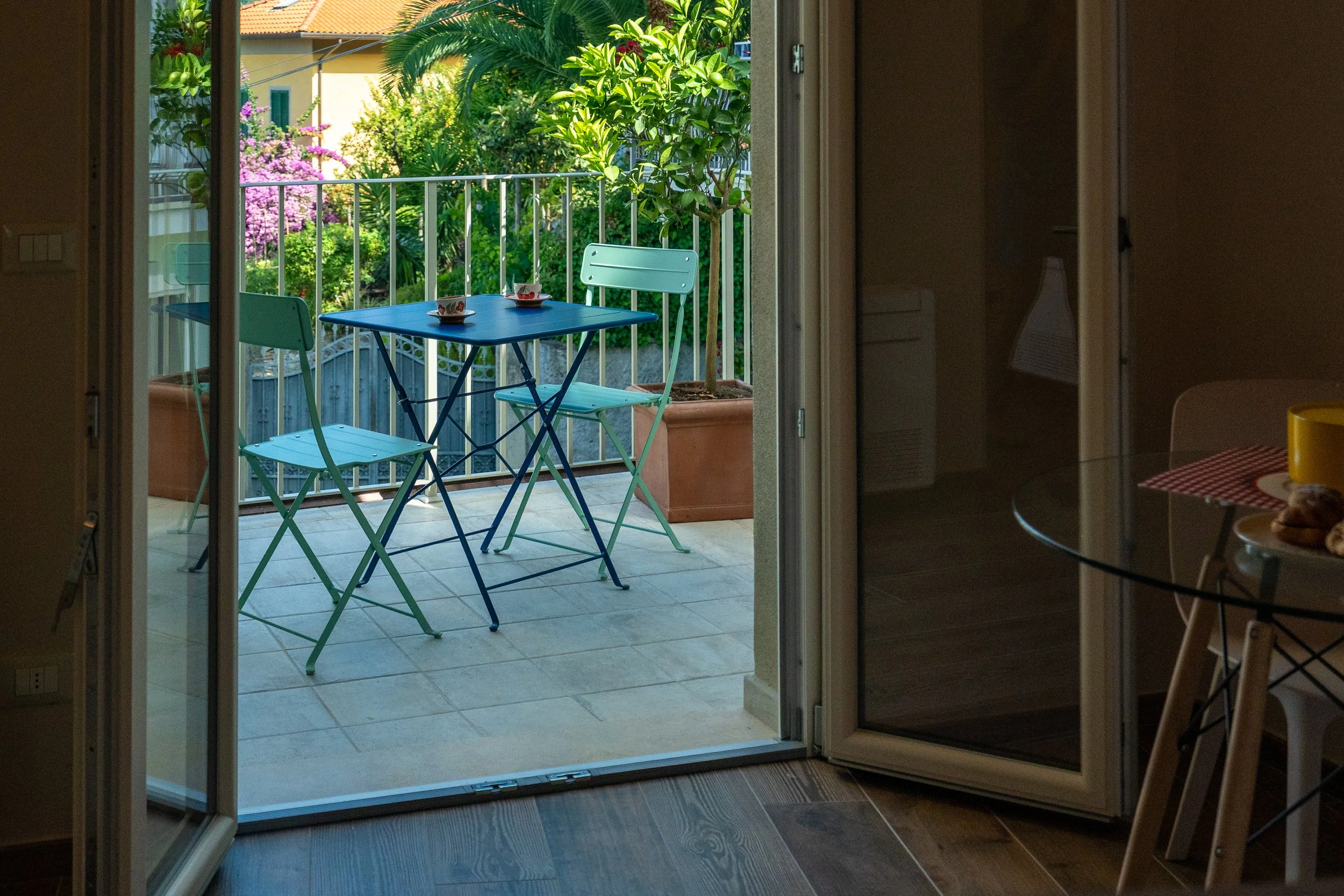 Vista di un balcone con un tavolo blu e due sedie di metallo, circondato da piante verdi e un albero in vaso, vista attraverso una porta scorrevole aperta da dentro una casa.  - Residenze Volpigiano