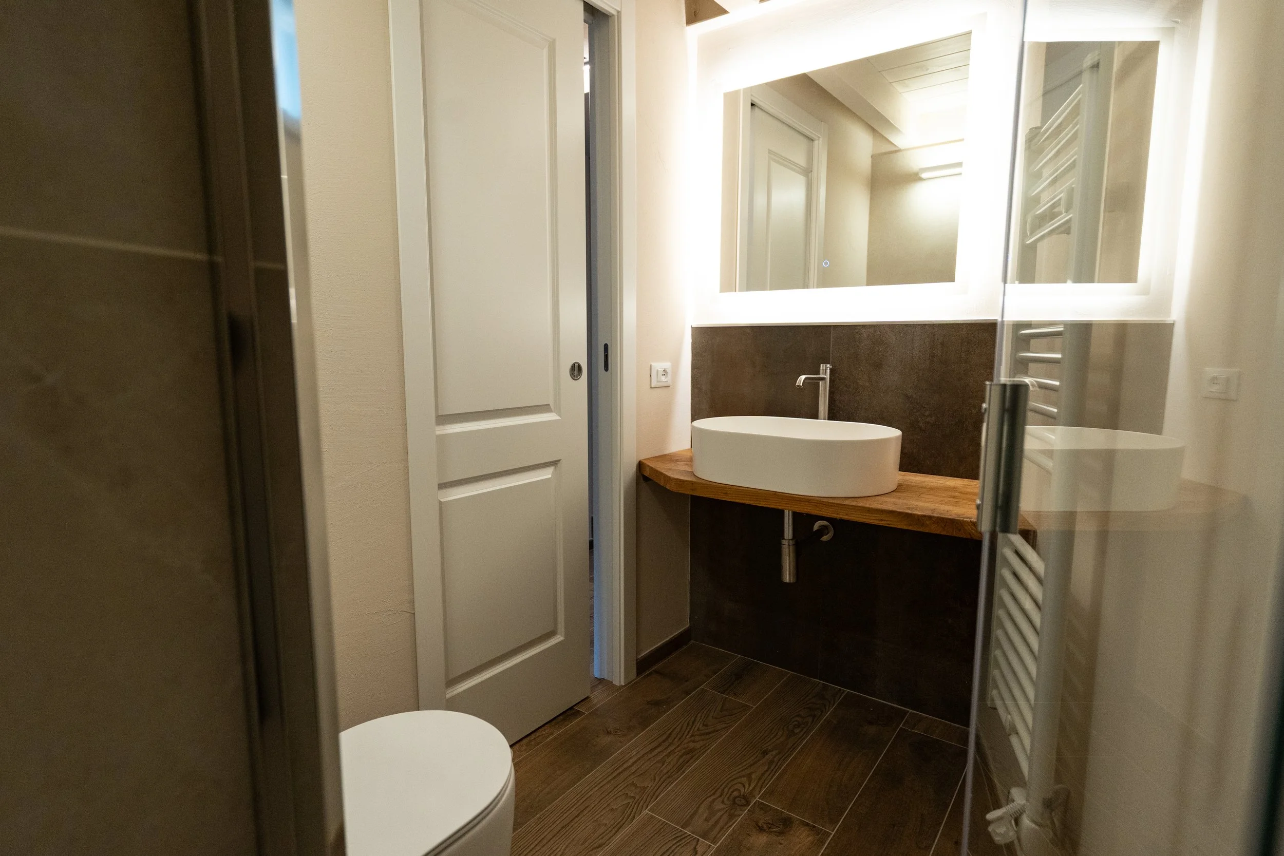Bagno moderno con lavabo rotondo bianco su banco di legno, grande specchio con illuminazione a LED, pareti in tonalità neutra, pavimento in legno scuro, porta scorrevole e termosifone bianco a raggio. - Residenze Volpigiano