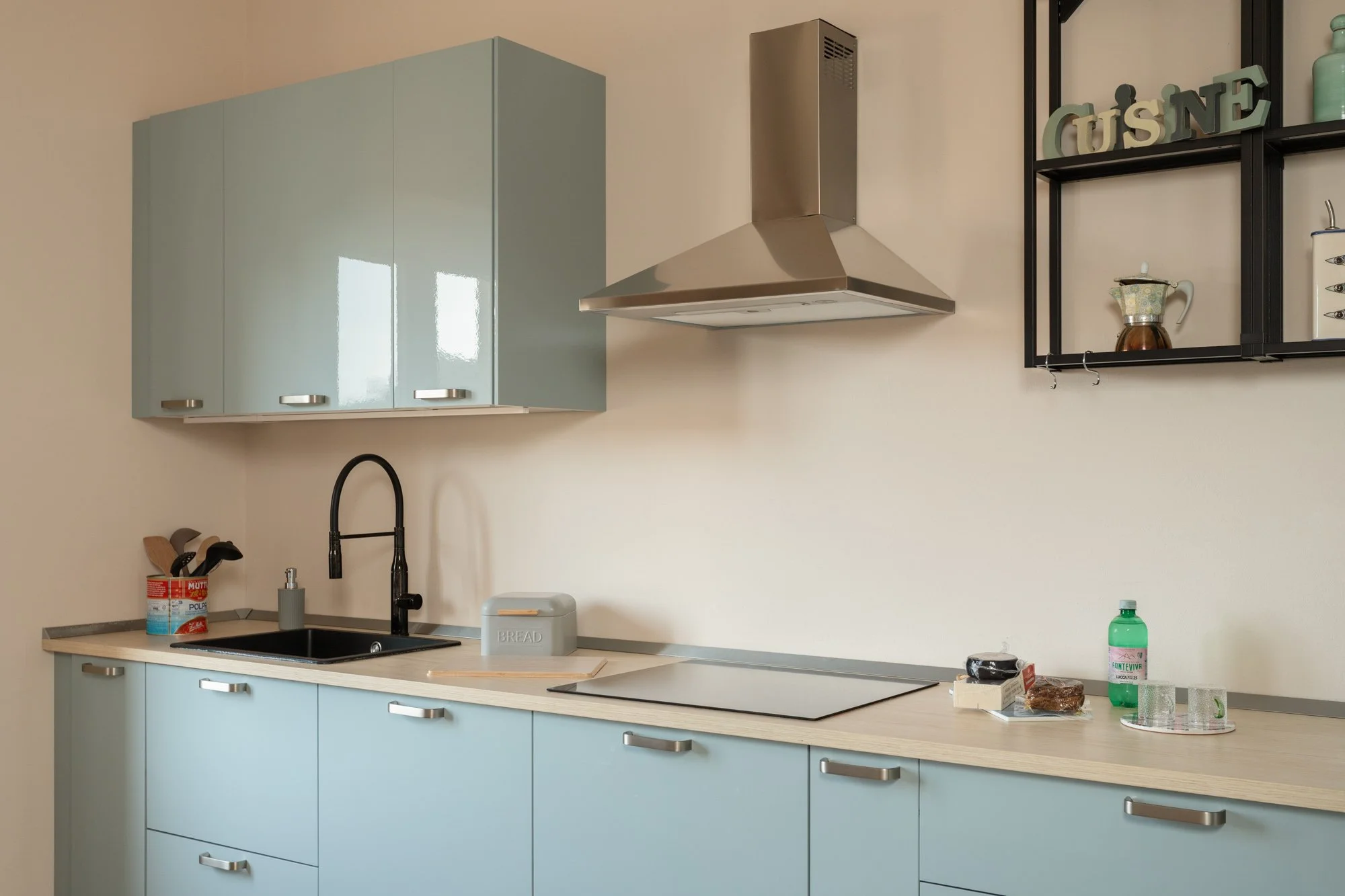 Cucina moderna con ante blu, piano in legno chiaro, zona lavello nera con rubinetto nero, cappa in acciaio, scaffale nero con decorazioni e oggetti da cucina, e alcuni alimenti e bevande sul piano.