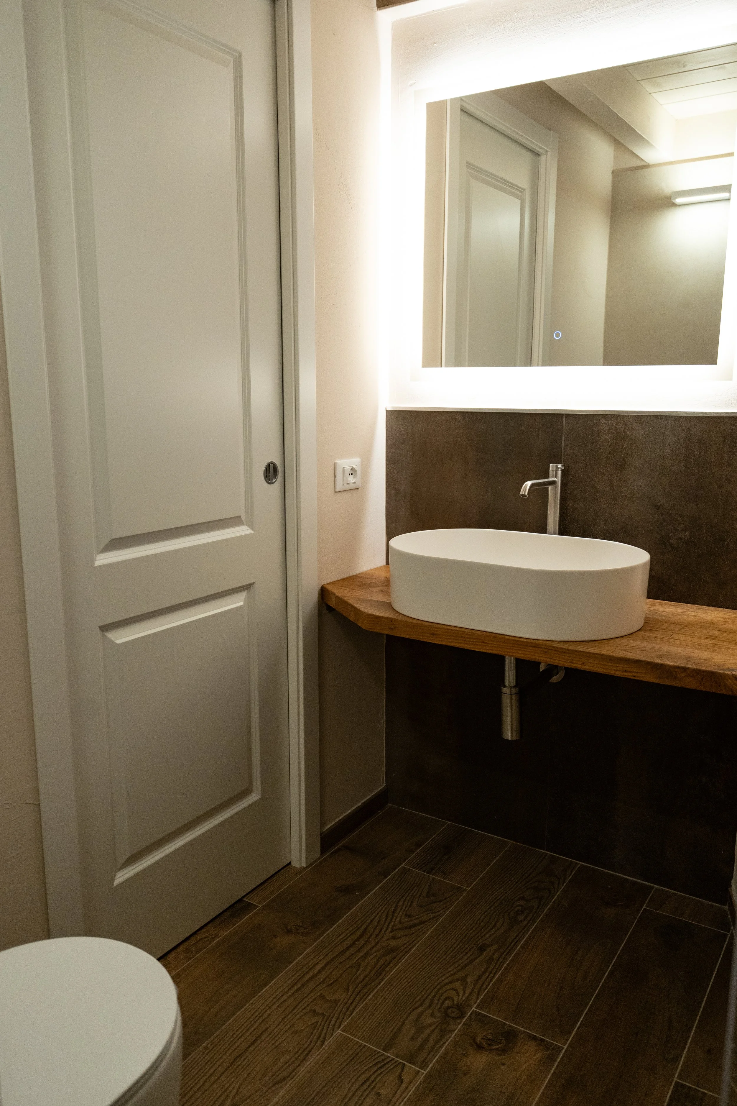 Bagno moderno con lavabo ovale bianco su piano di legno, grande specchio con cornice luminosa, pareti chiare e pavimento in legno scuro.  - Residenze Volpigiano