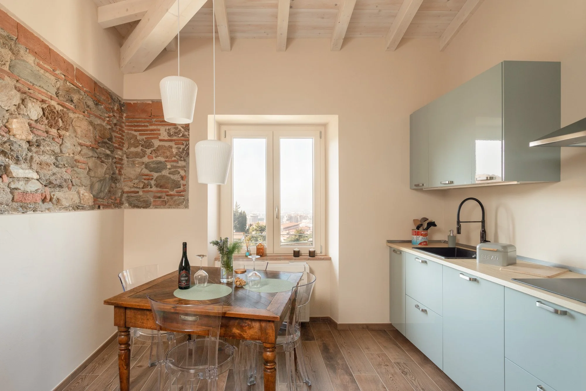 Interno di una cucina con parete di pietra, soffitto con travi di legno, tavolo di legno con sedie trasparenti, pensili azzurri, lavandino nero e finestra con vista sulla città.