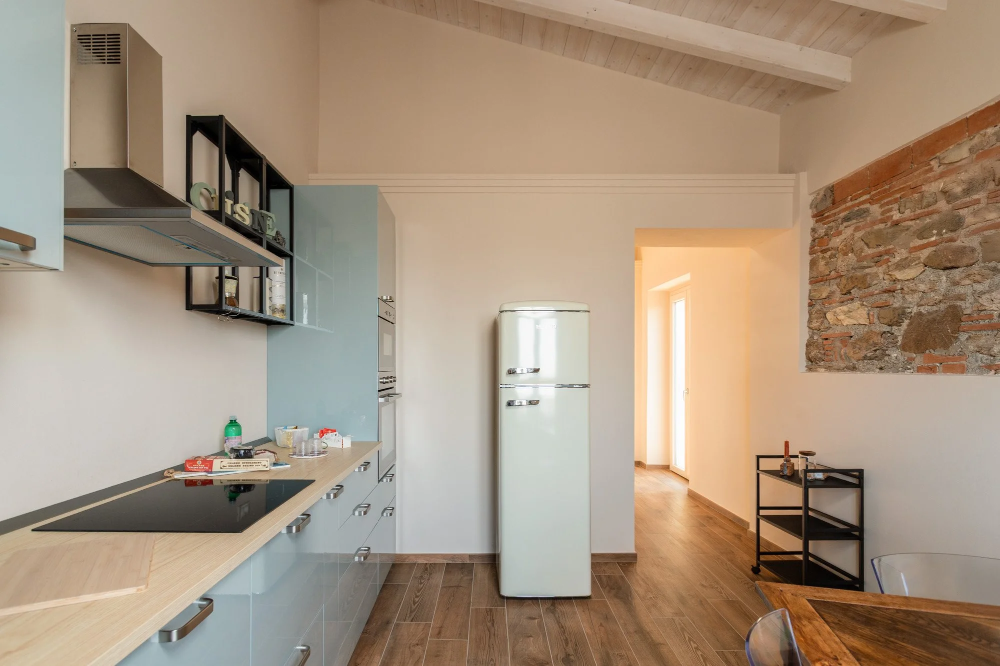 Cucina moderna con frigorifero bianco e piano cottura nero, pareti beige e soffitto con travi in legno, parete con mattoni a vista.