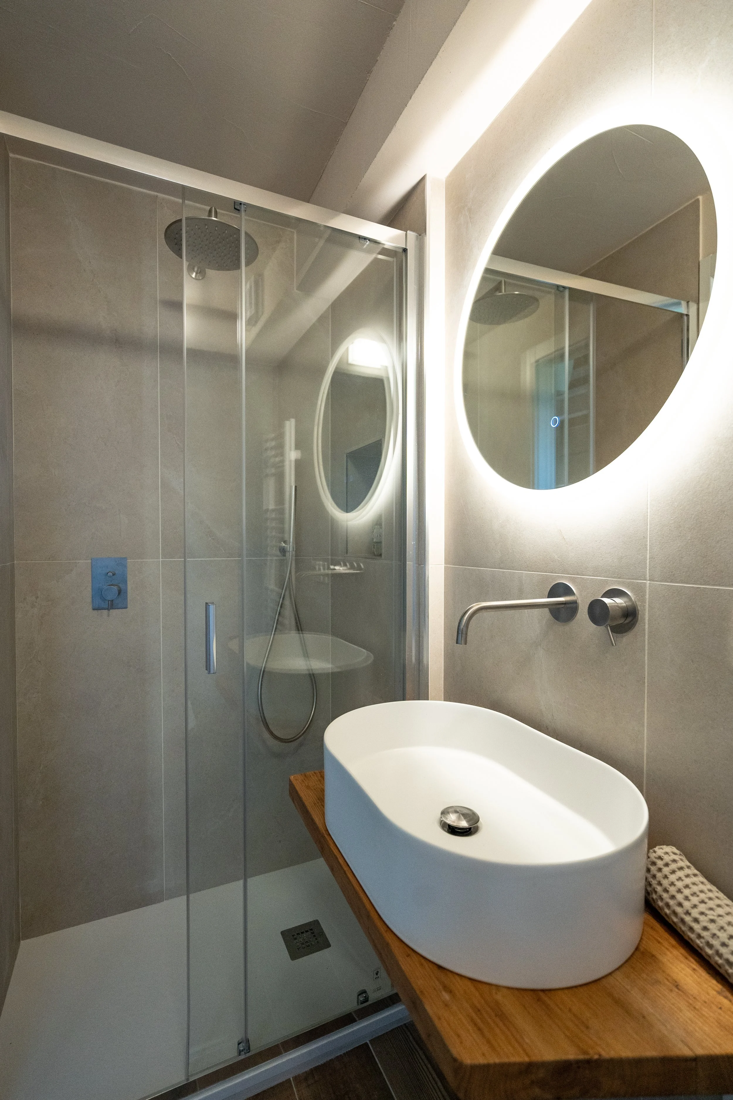 Bagno con lavabo bianco su panca di legno, grande specchio rotondo con illuminazione e doccia con porta di vetro. - Residenze Volpigiano