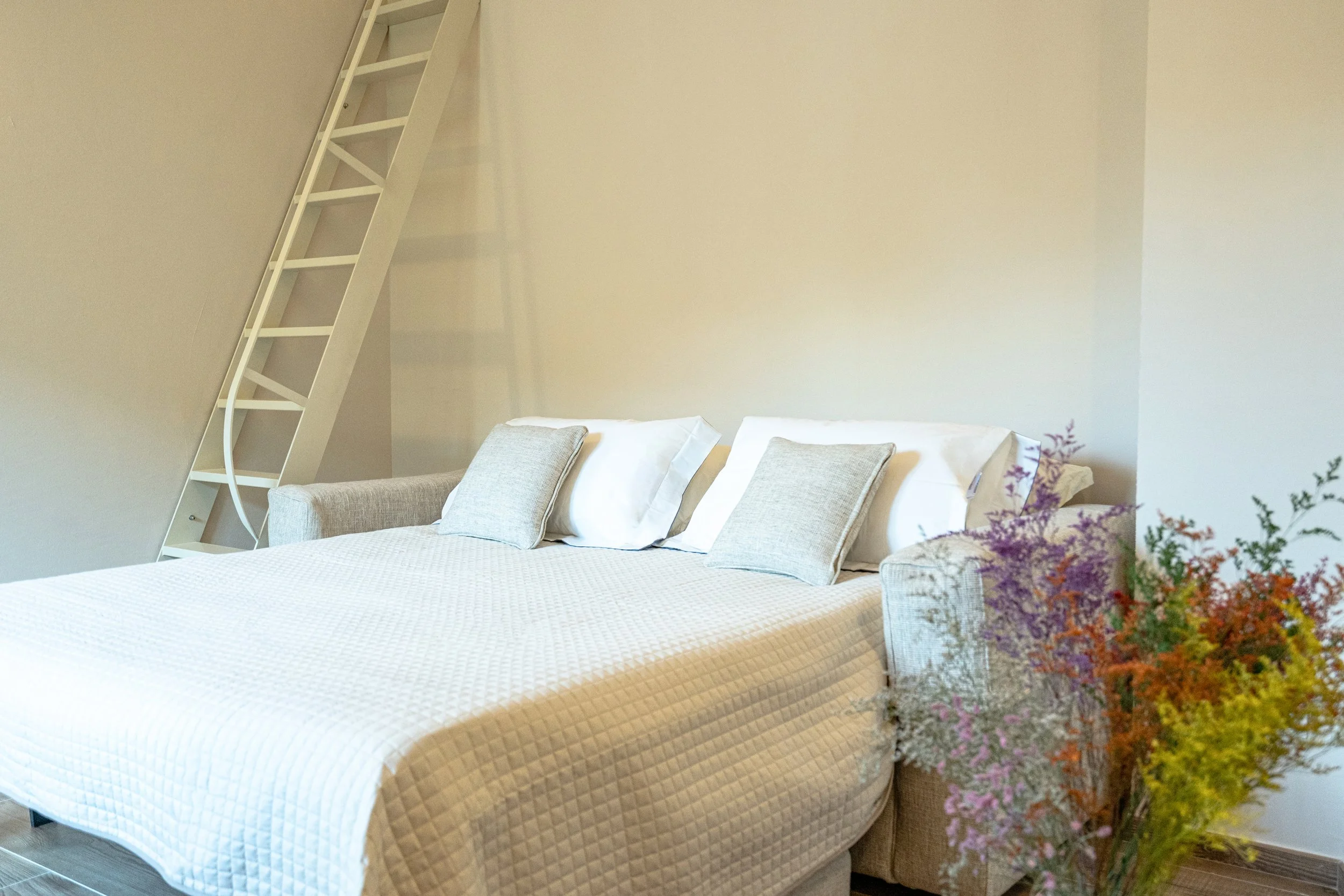 Letto matrimoniale con cuscini, accanto un vaso con fiori colorati, sullo sfondo parete bianca e una scala bianca di design. - Residenze Volpigiano