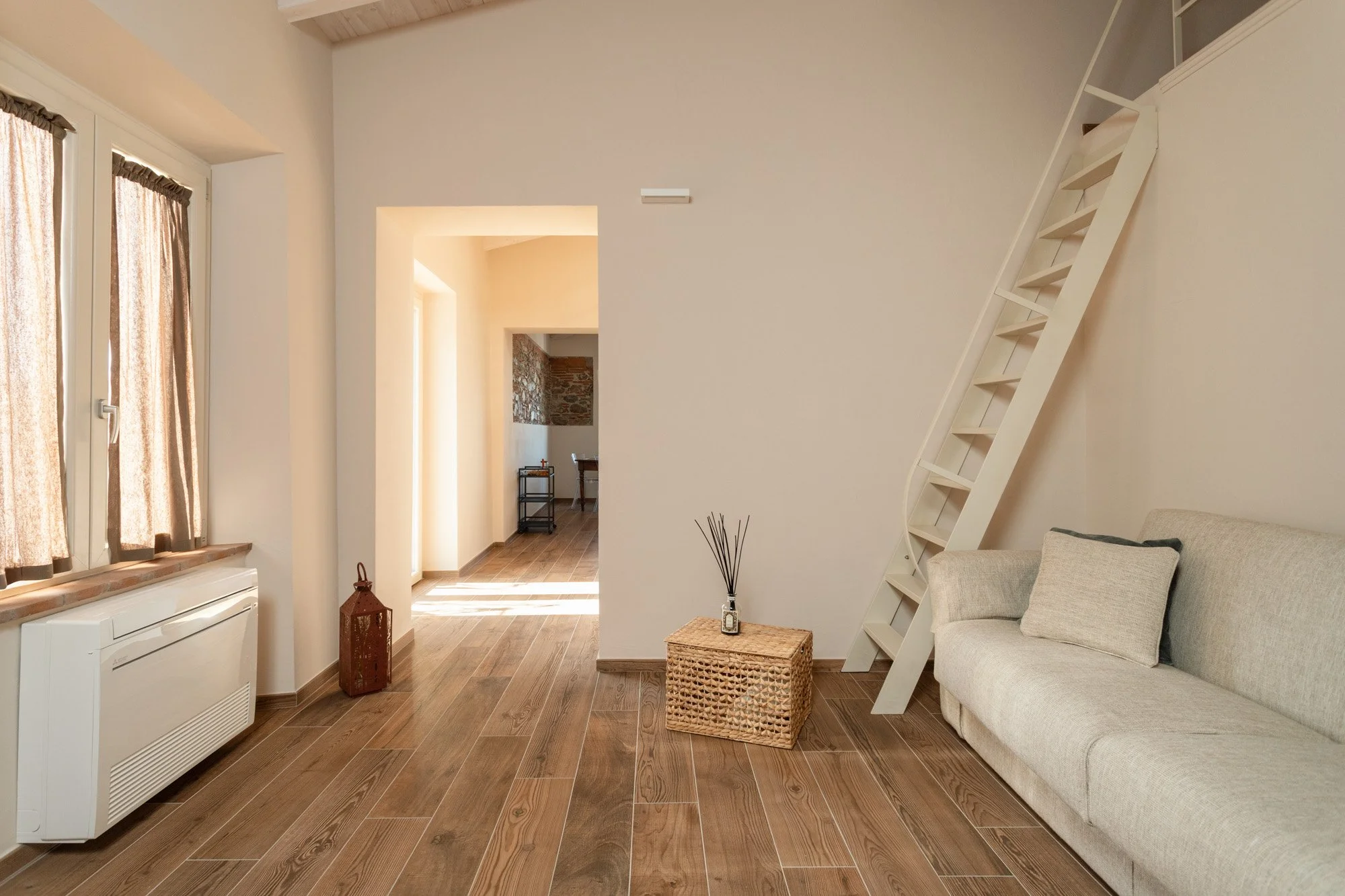 Interno di una stanza con pareti bianche, pavimento in legno, divano beige, scala a pioli bianca, finestra con tende beige, decorazioni minimaliste, porta che conduce a un altro spazio.