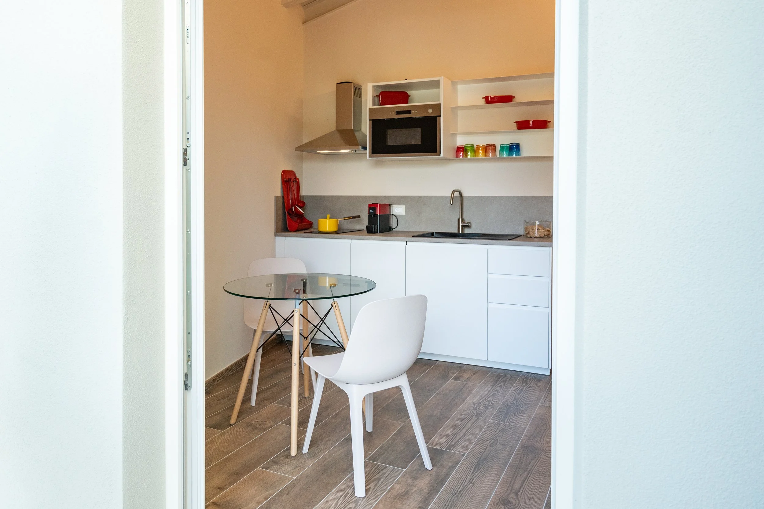Cucina moderna con mobili bianchi, tavolo in vetro con due sedie e scaffali con stoviglie colorate. - Residenze Volpigiano