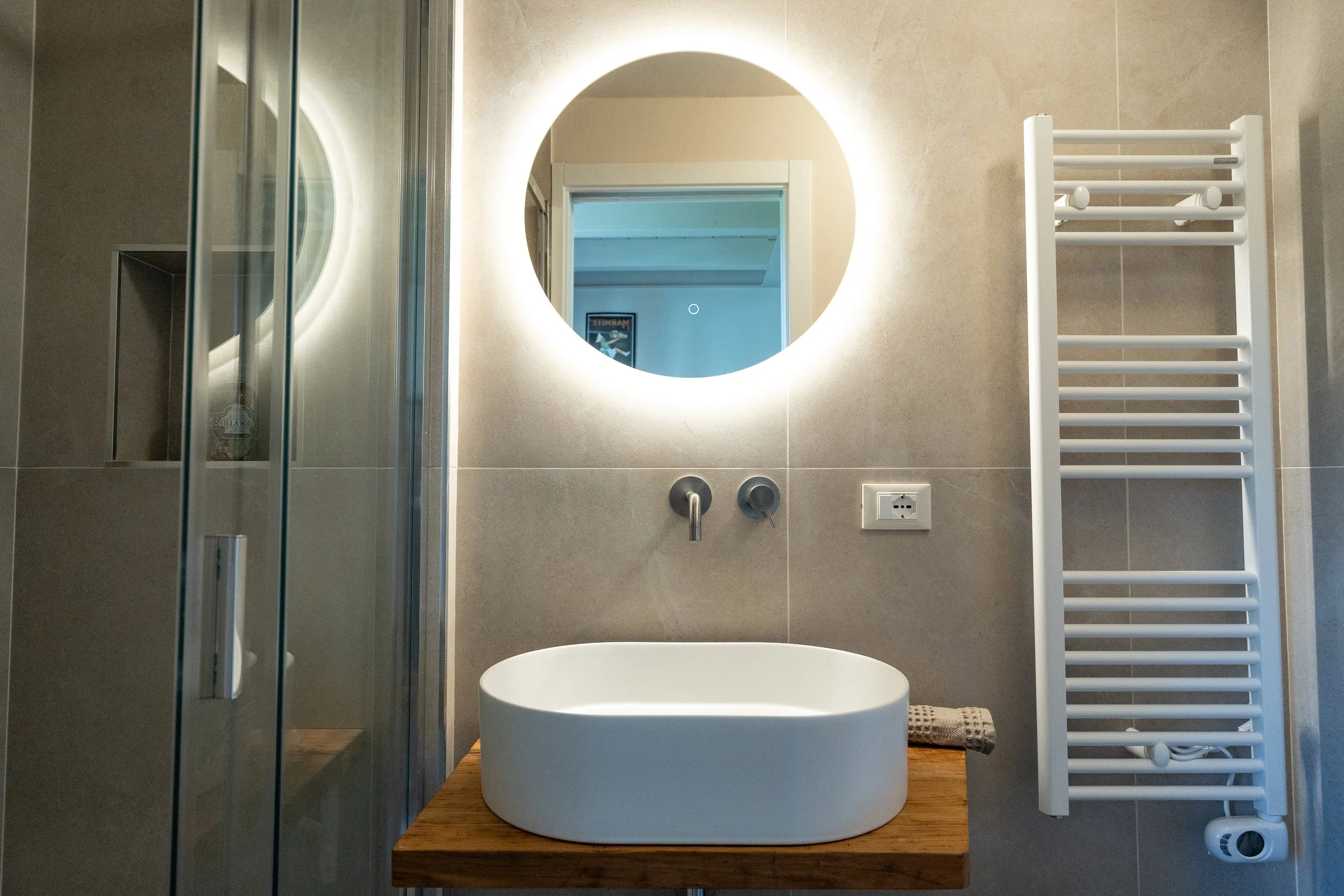 Specchio rotondo con illuminazione a LED sopra un lavabo bianco ovale su un mobile di legno in un bagno moderno - Residenze Volpigiano