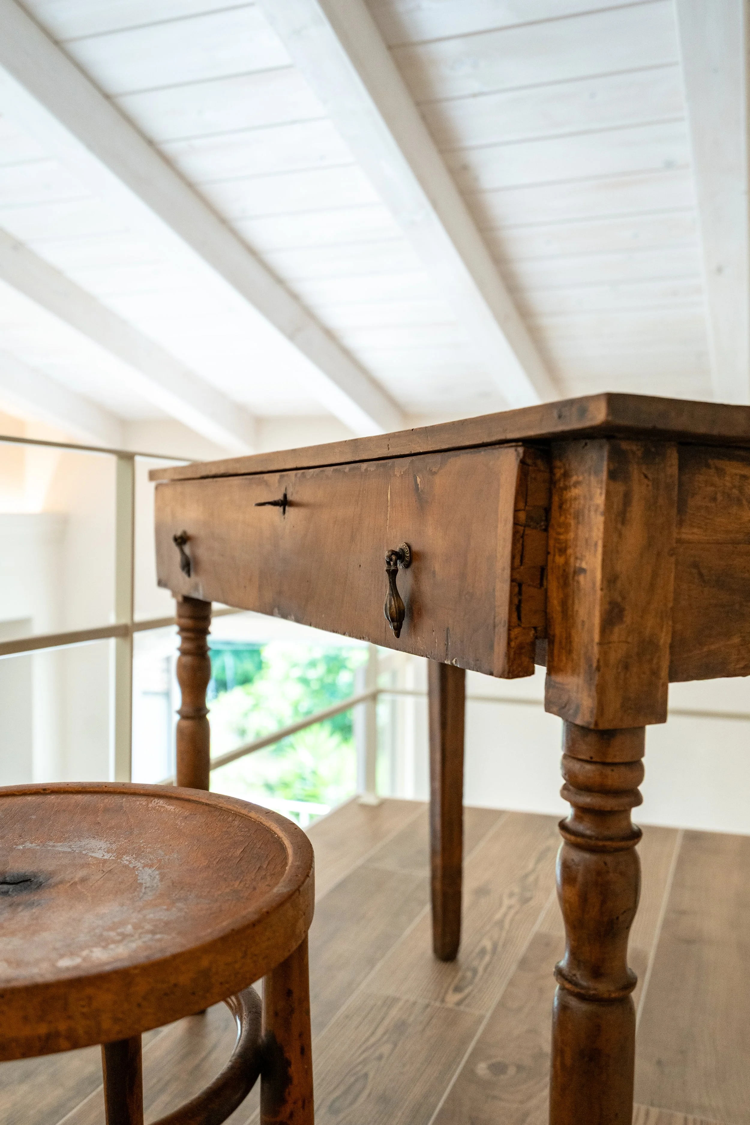 Una vecchia scrivania di legno rustico, posizionata accanto a uno sgabello di legno. Sullo sfondo, soffitto con travi di legno bianco e una ringhiera di sicurezza, in un ambiente luminoso con ampie finestre. - Residenze Volpigliano