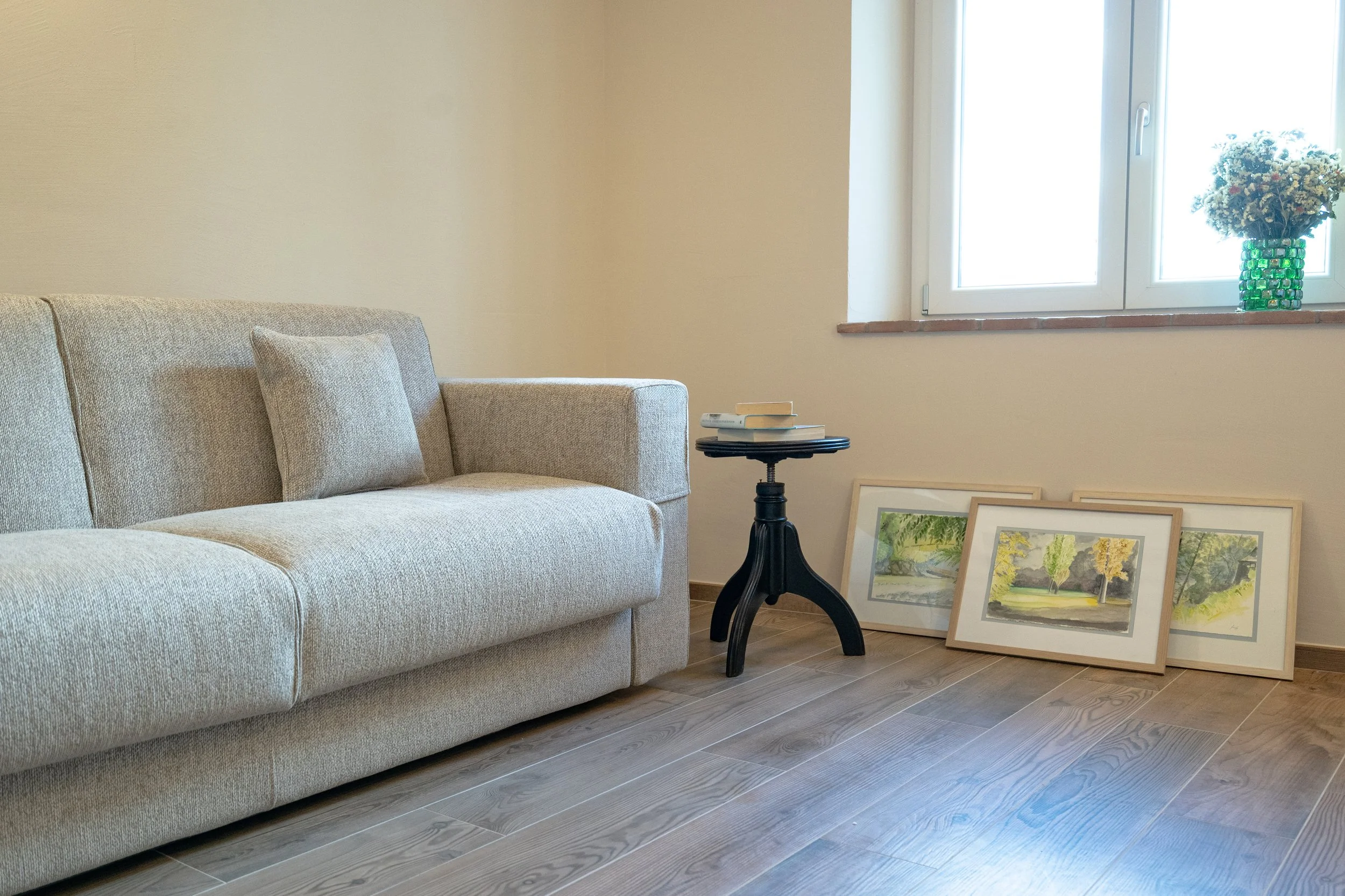 Soggiorno con divano beige, tavolino nero con libri, quadri appoggiati a terra, finestra con vasi di fiori - Residenze Volpigiano