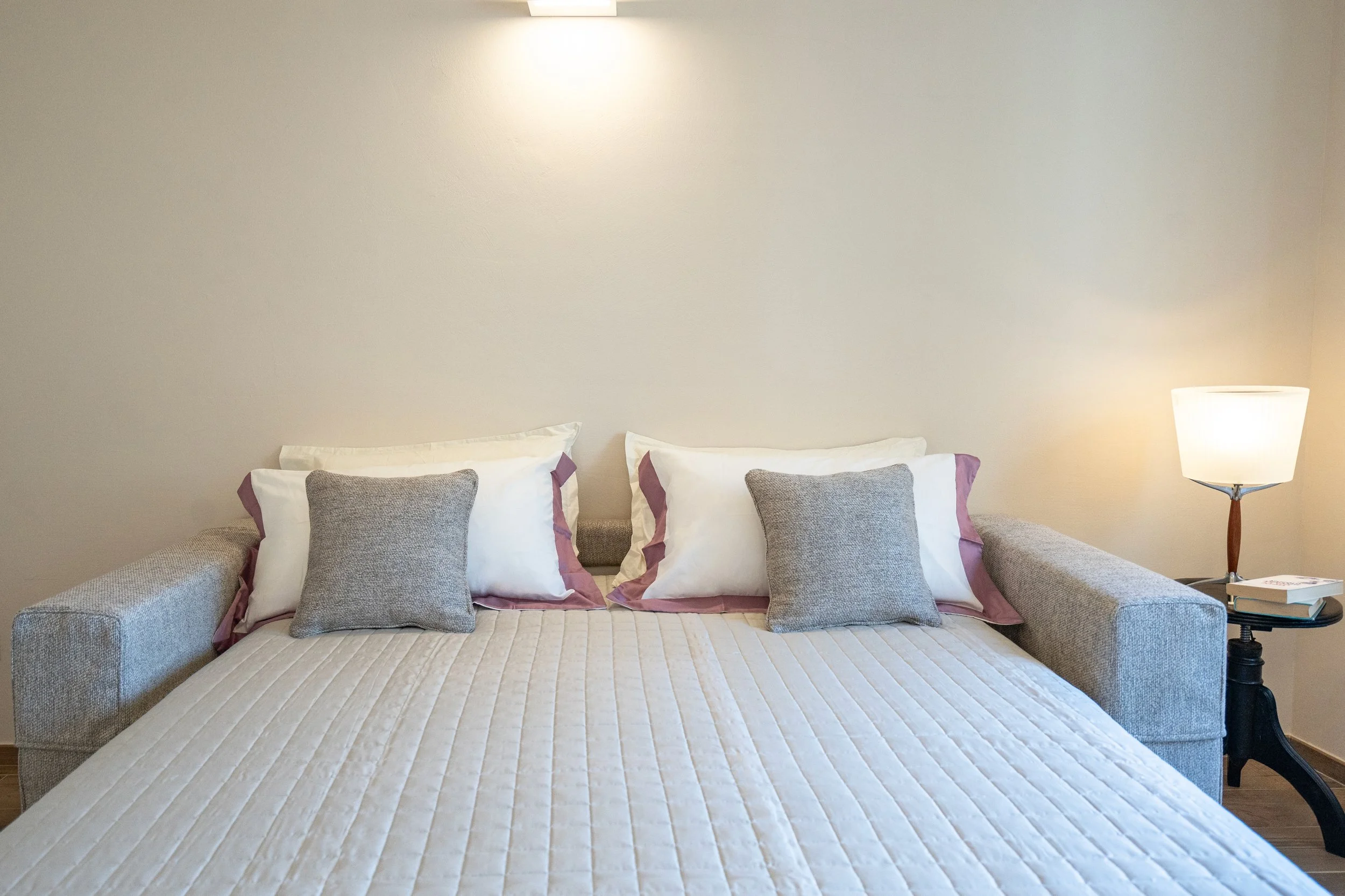 Letto matrimoniale con cuscini grigi, coperte bianche e divano grigio ai lati, lampada accesa su un tavolino nero con libri accanto, parete beige sullo sfondo. - Residenze Volpigiano