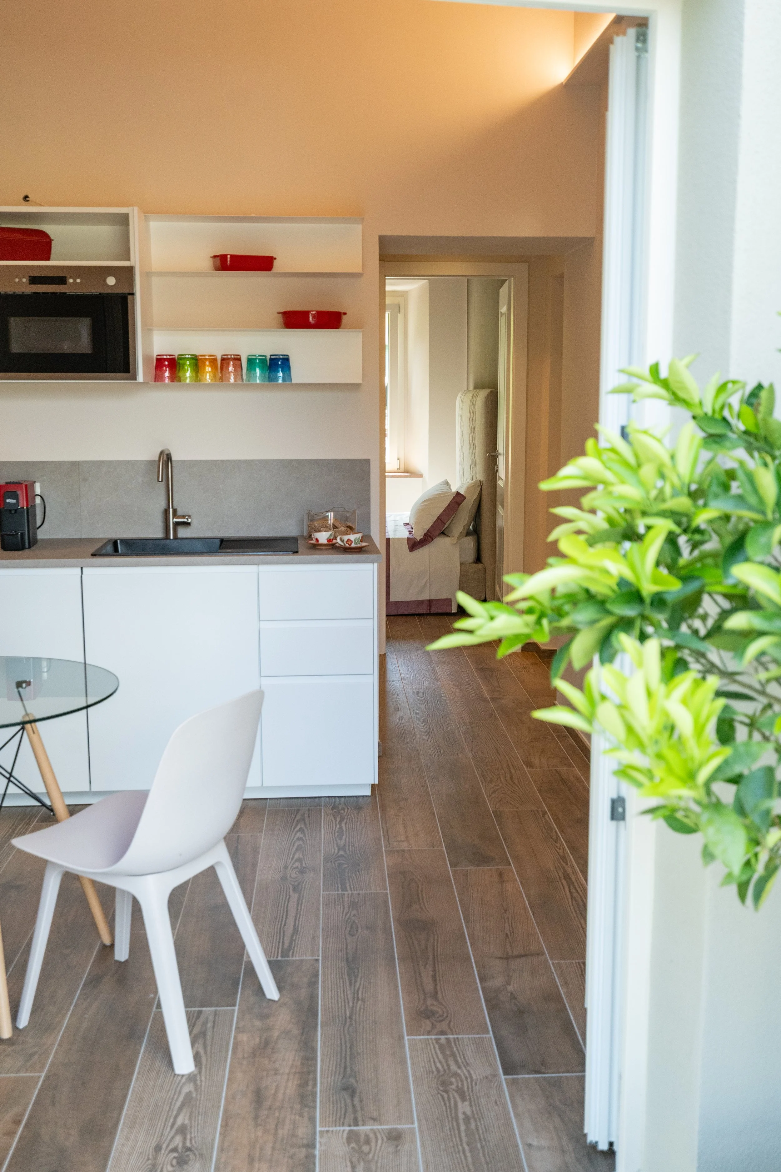 Interno di una cucina con mobili bianchi, piano di lavoro grigio, e accessori colorati come bicchieri e ciotole. Sul tavolo di vetro c'è una sedia bianca, e sul lato destro un albero con foglie verdi.  - Residenze Volpigiano