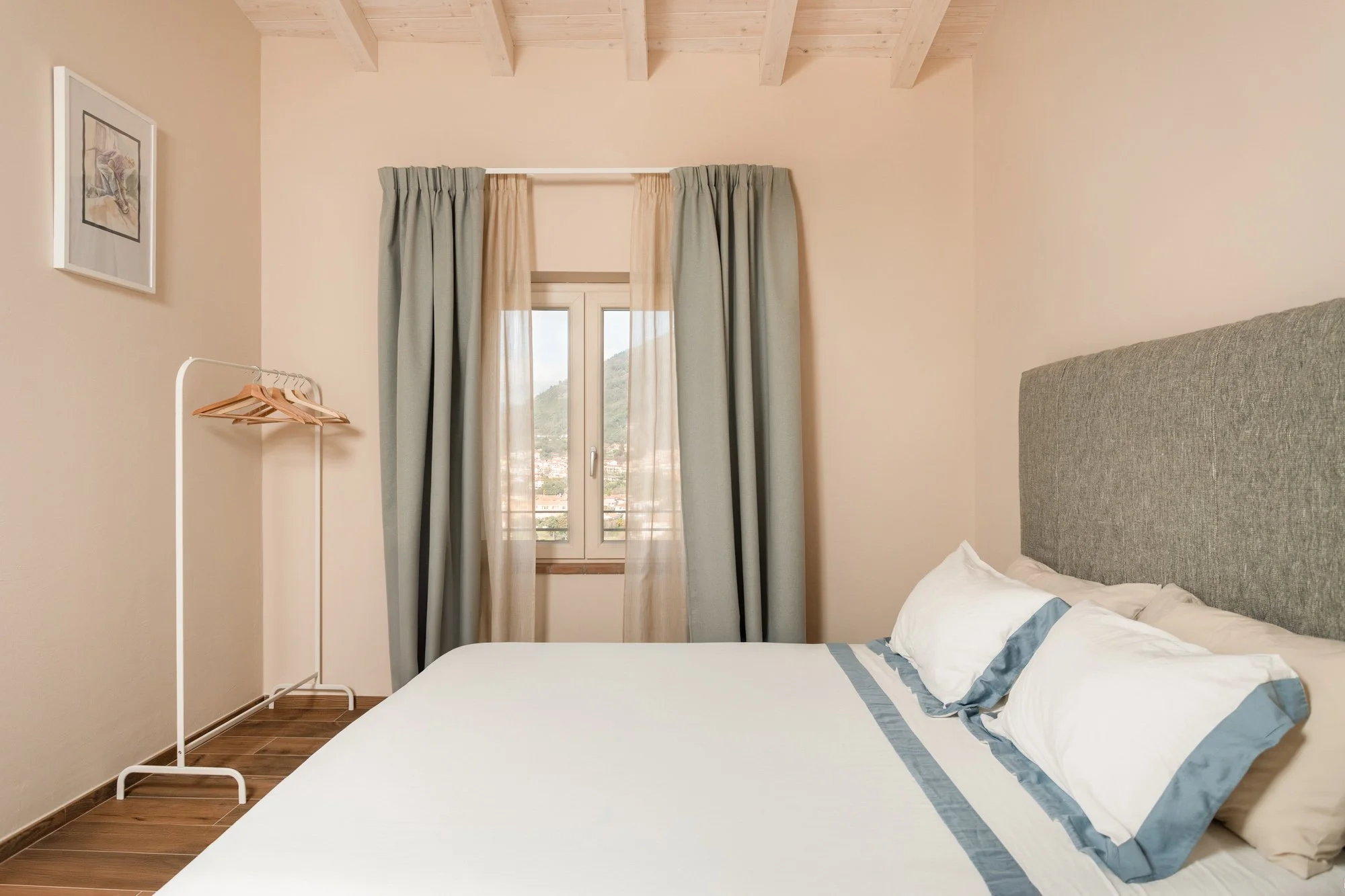 Una camera da letto con un letto con testiera grigia, lenzuola bianche con dettagli blu, due cuscini, una finestra con tende verdi e beige, pareti beige, pavimento in parquet e un quadro appeso alla parete.