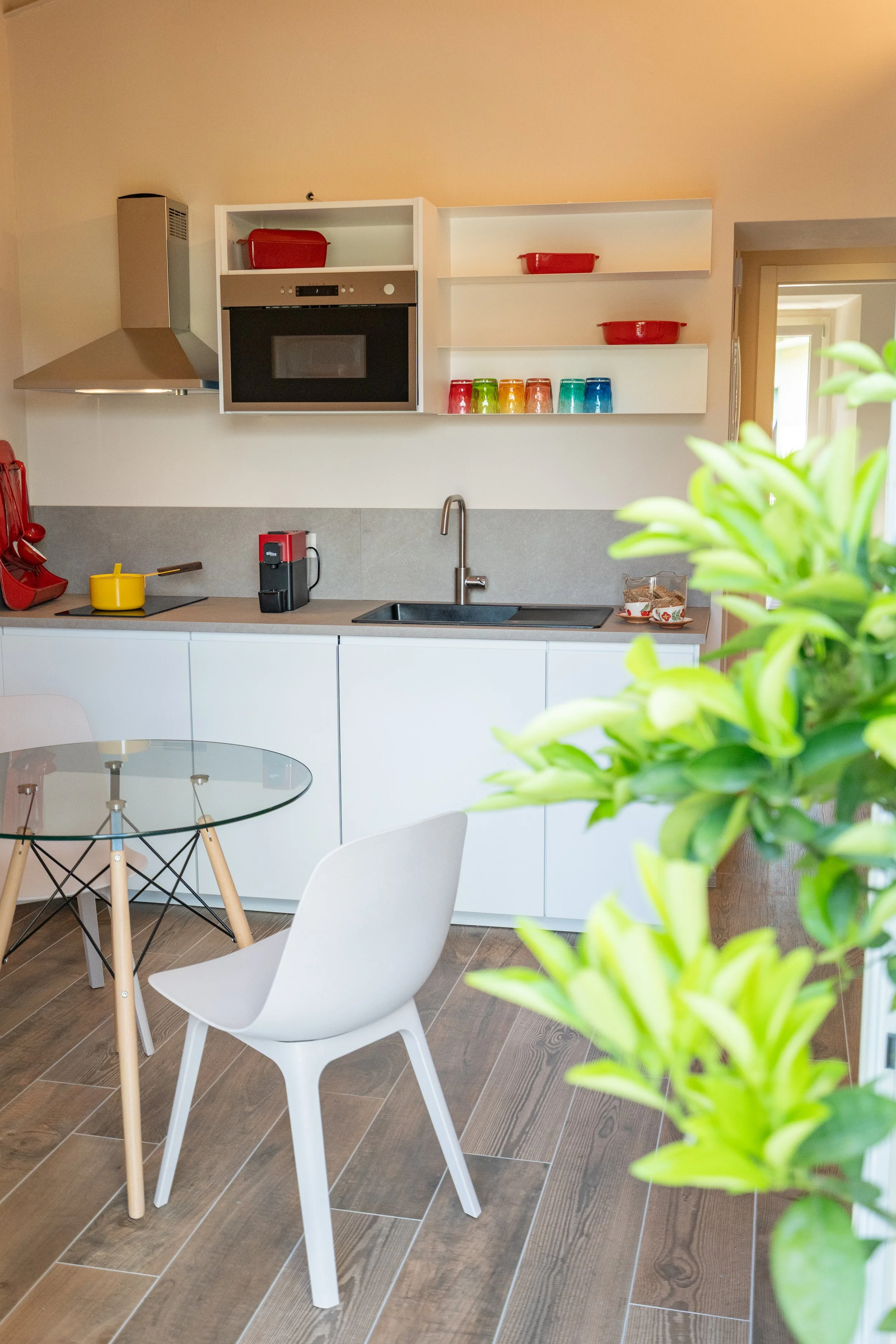 Cucina moderna con armadietti bianchi, piano di lavoro grigio, fornello elettrico, microonde nero, macchina da caffè, scaffali con piatti colorati e ciotole rosse, sedia bianca, tavolino rotondo con gambe in legno e sedie - Residenze Volpigliano