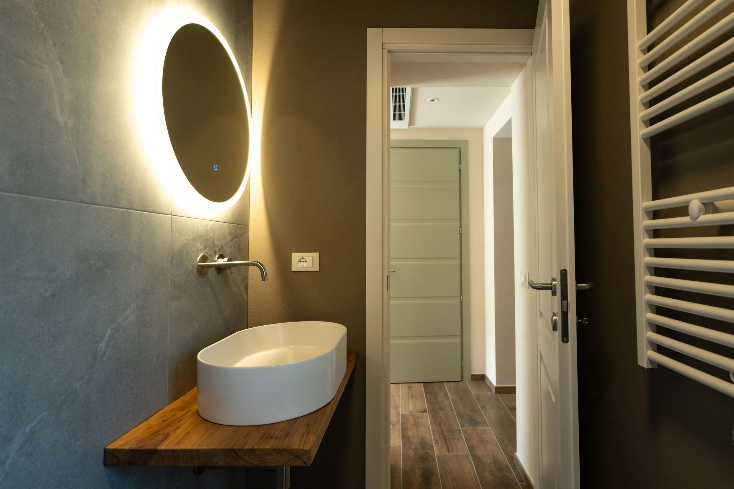 Bagno con lavabo su top in legno, specchio rotondo con luci a LED, porta aperta che mostra un corridoio, pavimento in piastrelle in stile legno, termosalviette bianco a parete. - Residenze Volpigiano