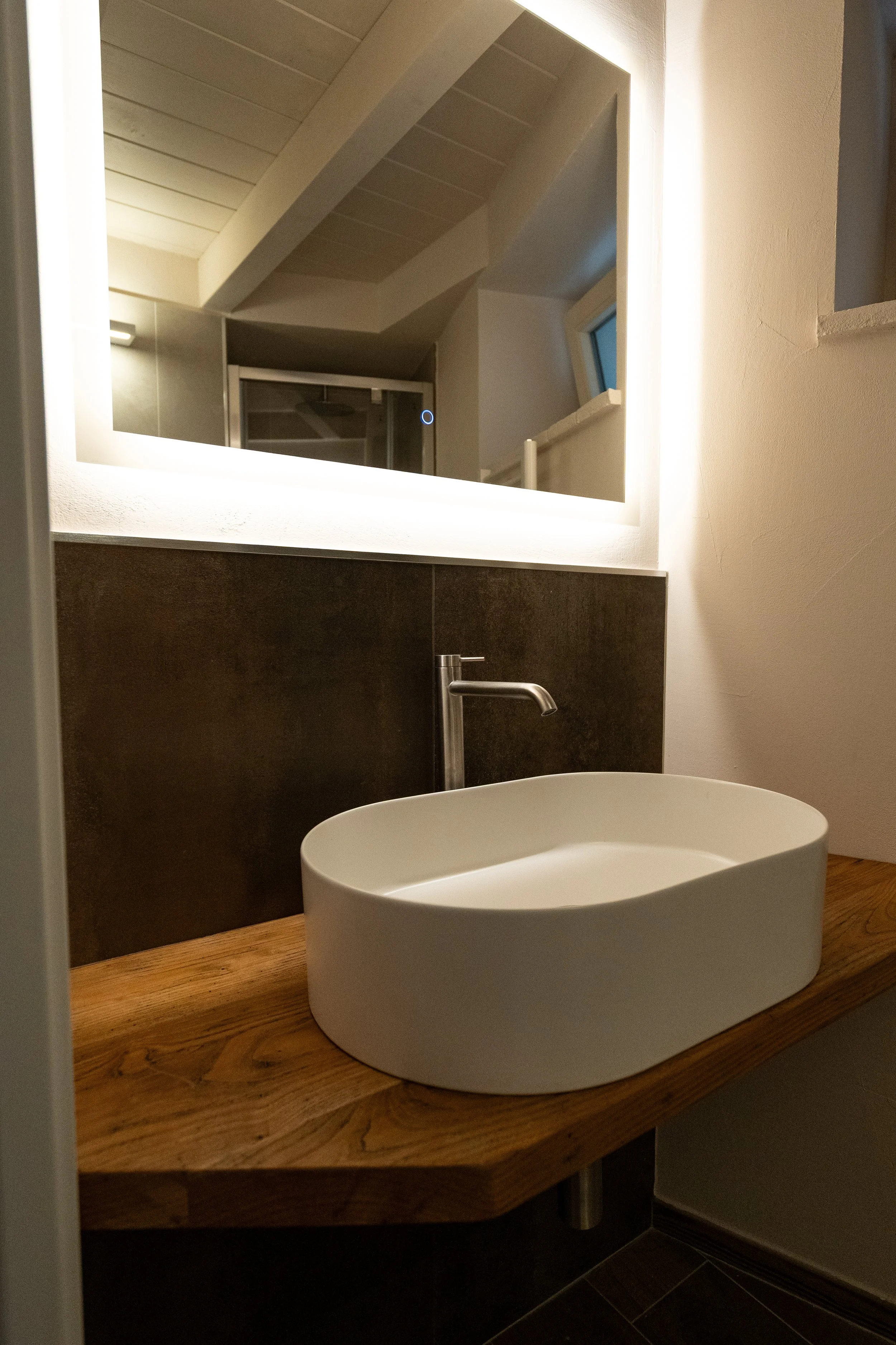 Lavabo da tavolo in ceramica bianca su mensola di legno, con specchio con illuminazione integrata e finestra in una stanza moderna. - Residenze Volpigiano