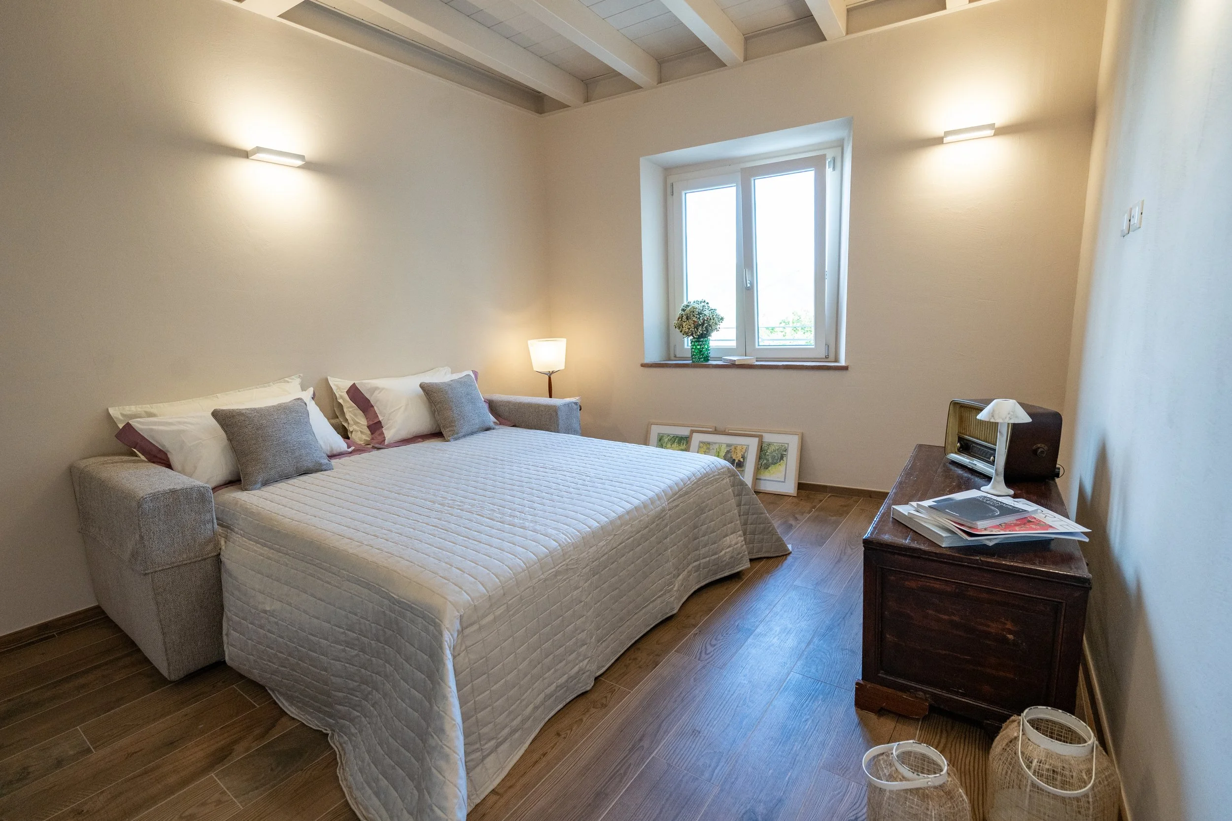 Camera da letto con letto matrimoniale, cuscini color grigio e bianco, finestra con fiori in vaso, lampade da parete e tavolo con oggetti decorativi. - Residenze Volpigiano