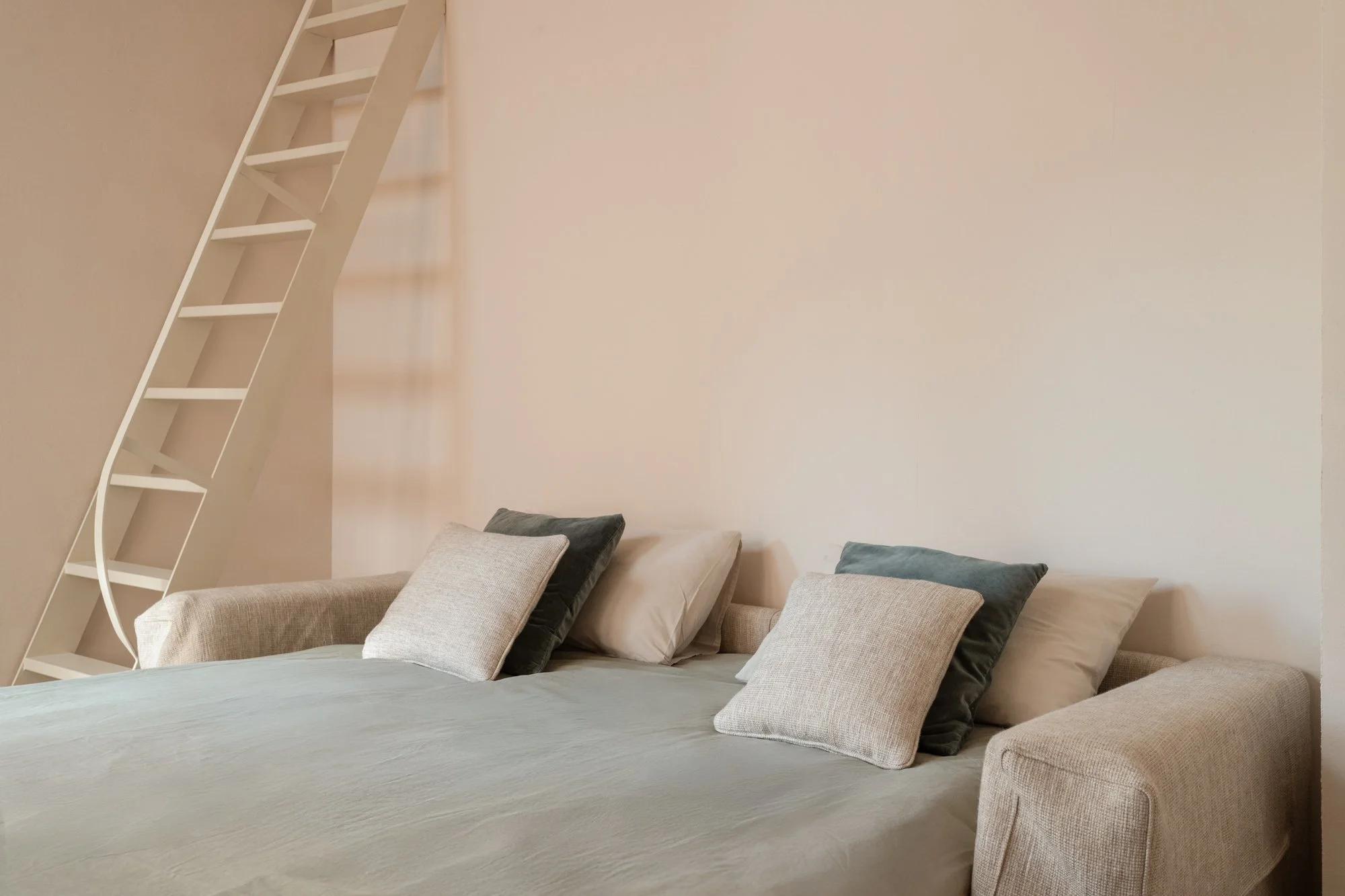 Divano beige con cuscini grigi e blu, pareti bianche e una scaletta bianca con rampe curve in un soggiorno moderno.