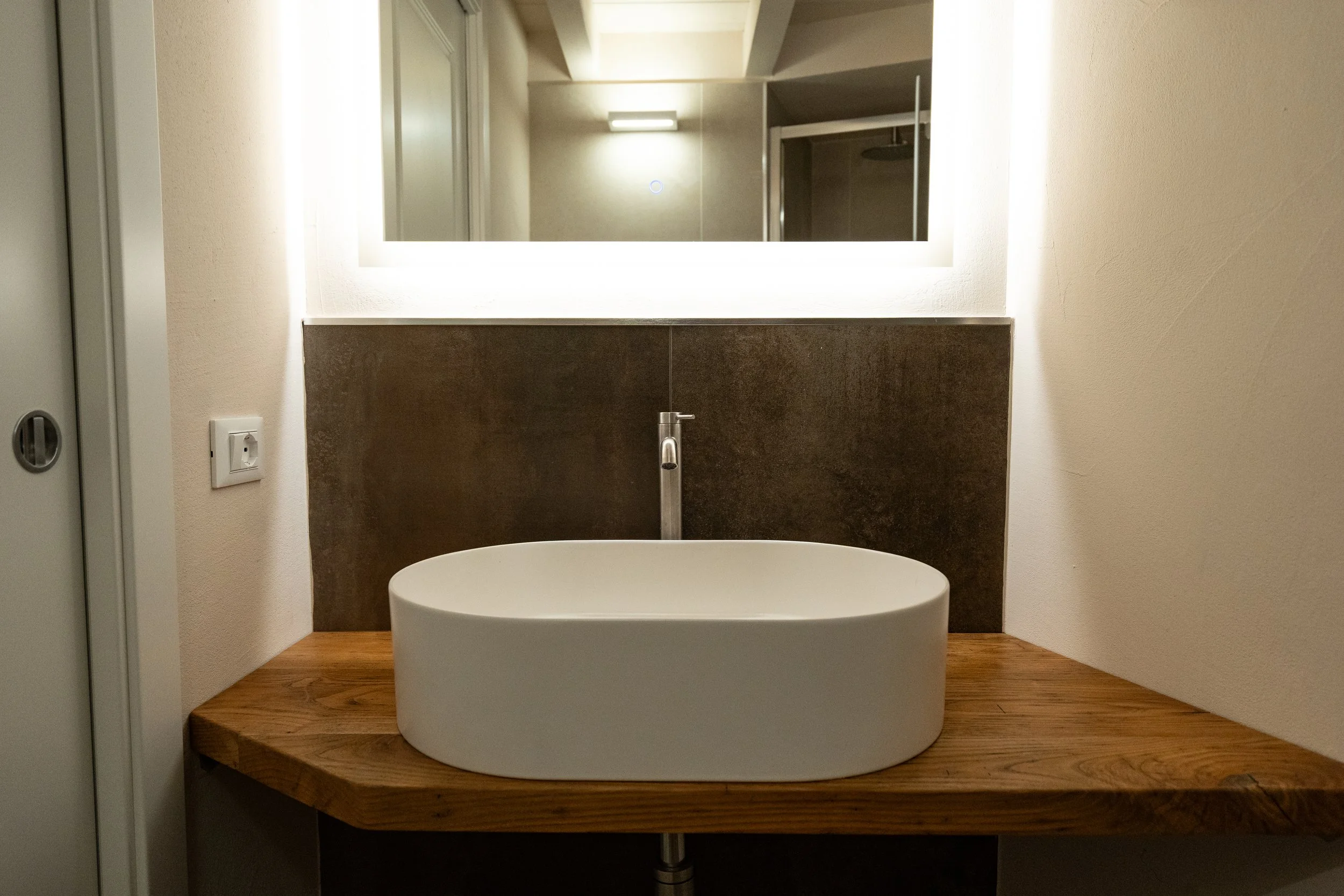 Lavandino rotondo bianco su un piano di legno in un bagno moderno con specchiera luminosa. - Residenze Volpigiano